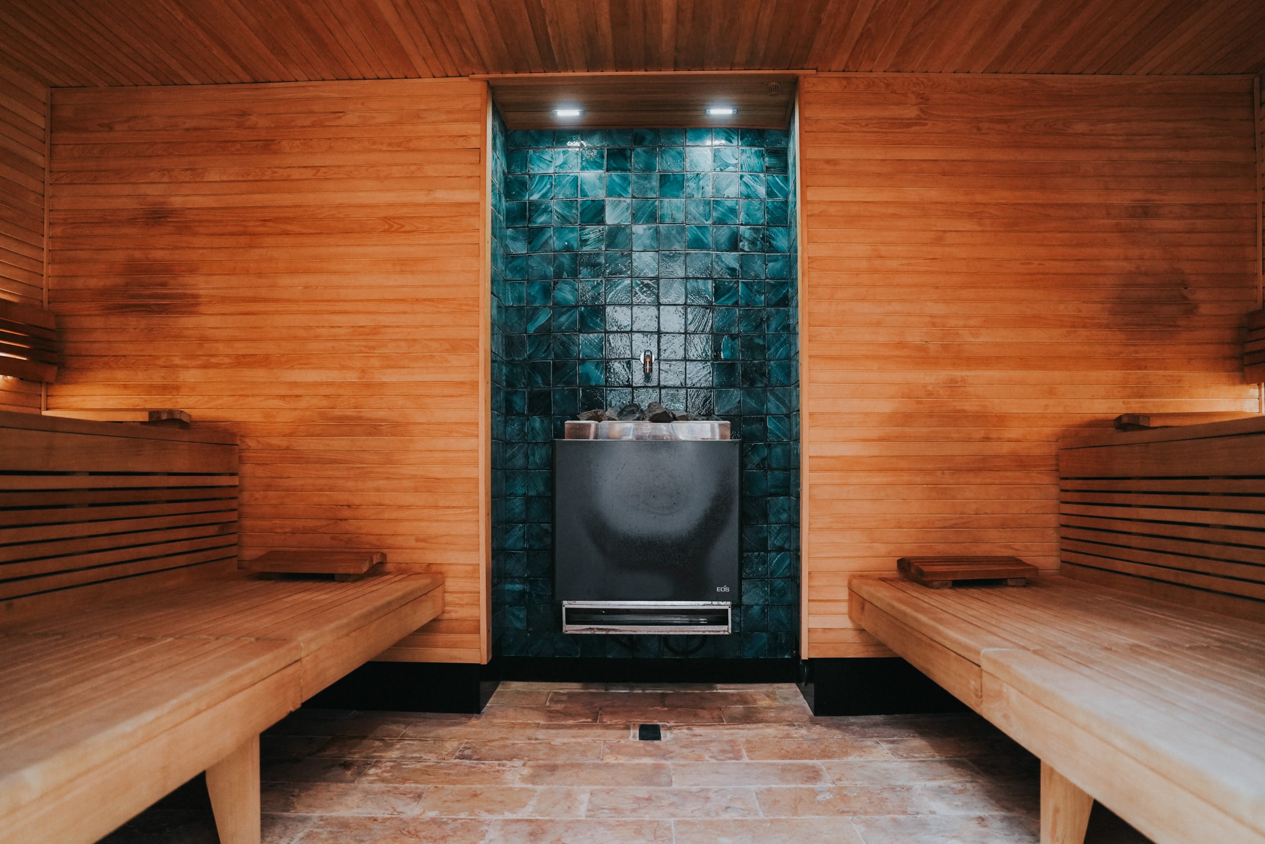 nterior of a wooden sauna room with tiered bench seating surrounding a central heater against a dark blue tiled wall accent.