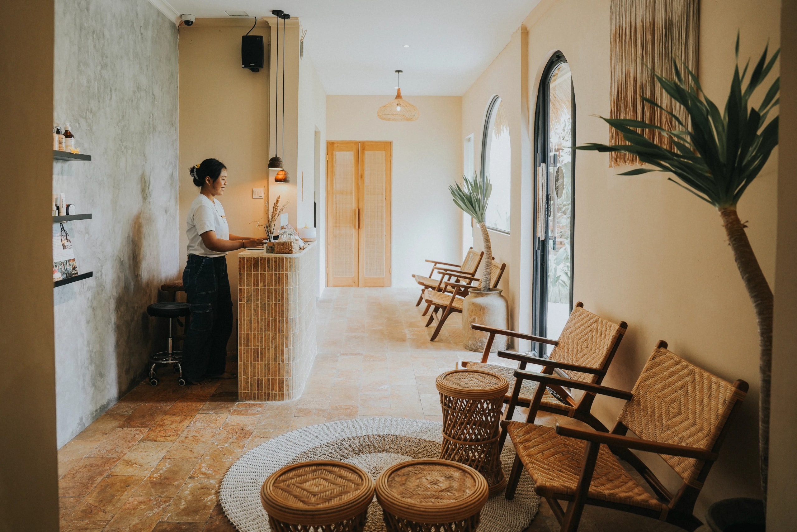 Bright spa reception area with a tiled front desk, receptionist, woven seating, neutral tones, and tropical potted plants.
