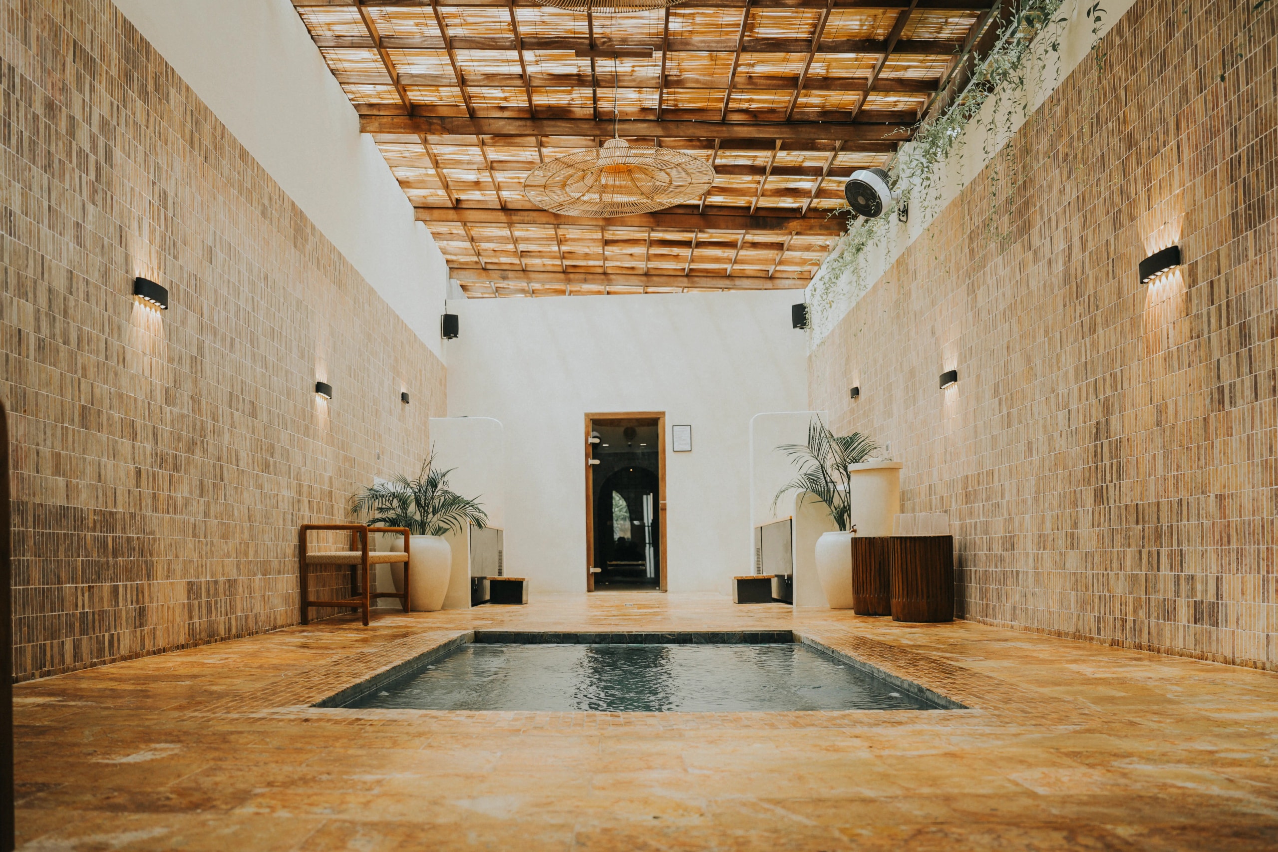 Spacious indoor wellness area featuring tiled walls, a wooden slat ceiling, wall sconces, plant accents, and a central plunge pool.