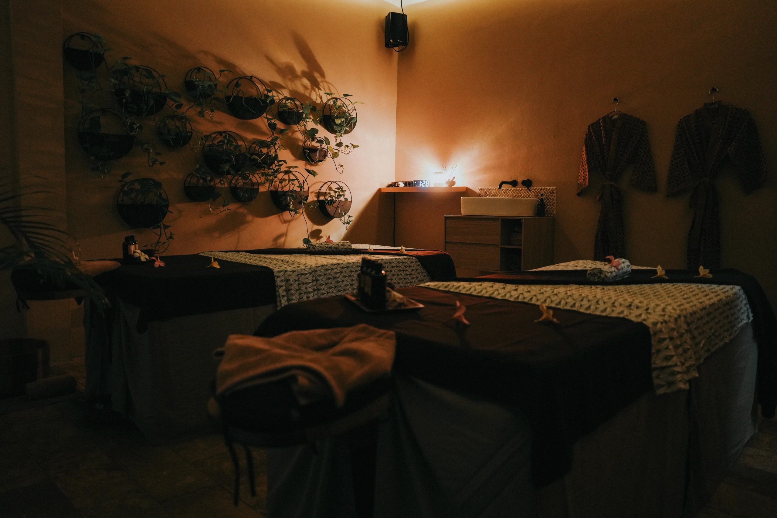 Dimly lit couple's massage room featuring two treatment tables, warm mood lighting, hanging bathrobes, and a wall adorned with potted plants.