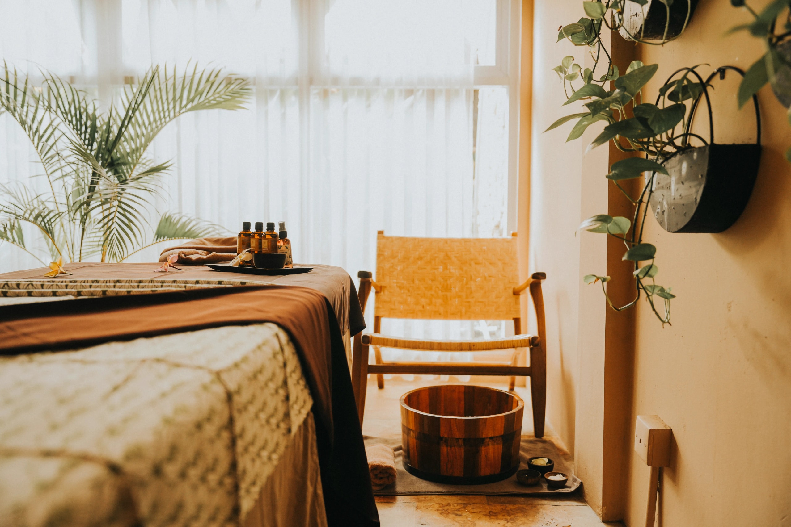 Spa room corner showing a prepared massage table, a wooden foot bath basin on the floor, and a woven armchair near a window with sheer curtains.