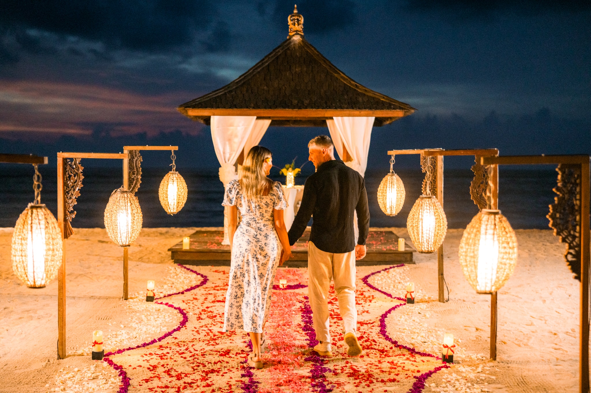 A couple holding hands as they walk down a beach pathway lined with flower petals and lanterns toward a glowing beachfront gazebo at dusk.