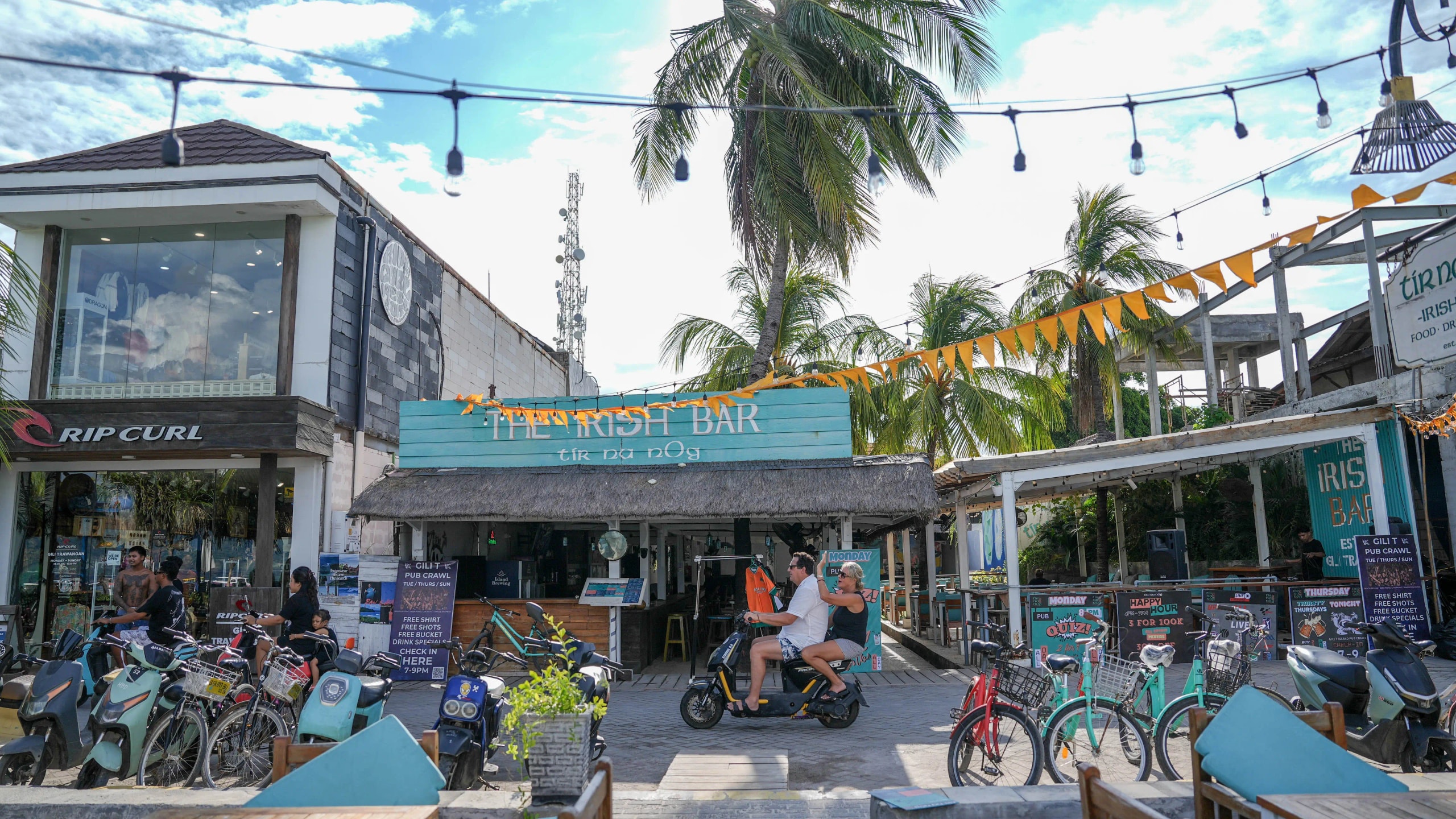 A bustling tropical street scene featuring "The Irish Bar," parked bicycles, scooters, and tourists riding a moped past a Rip Curl store.