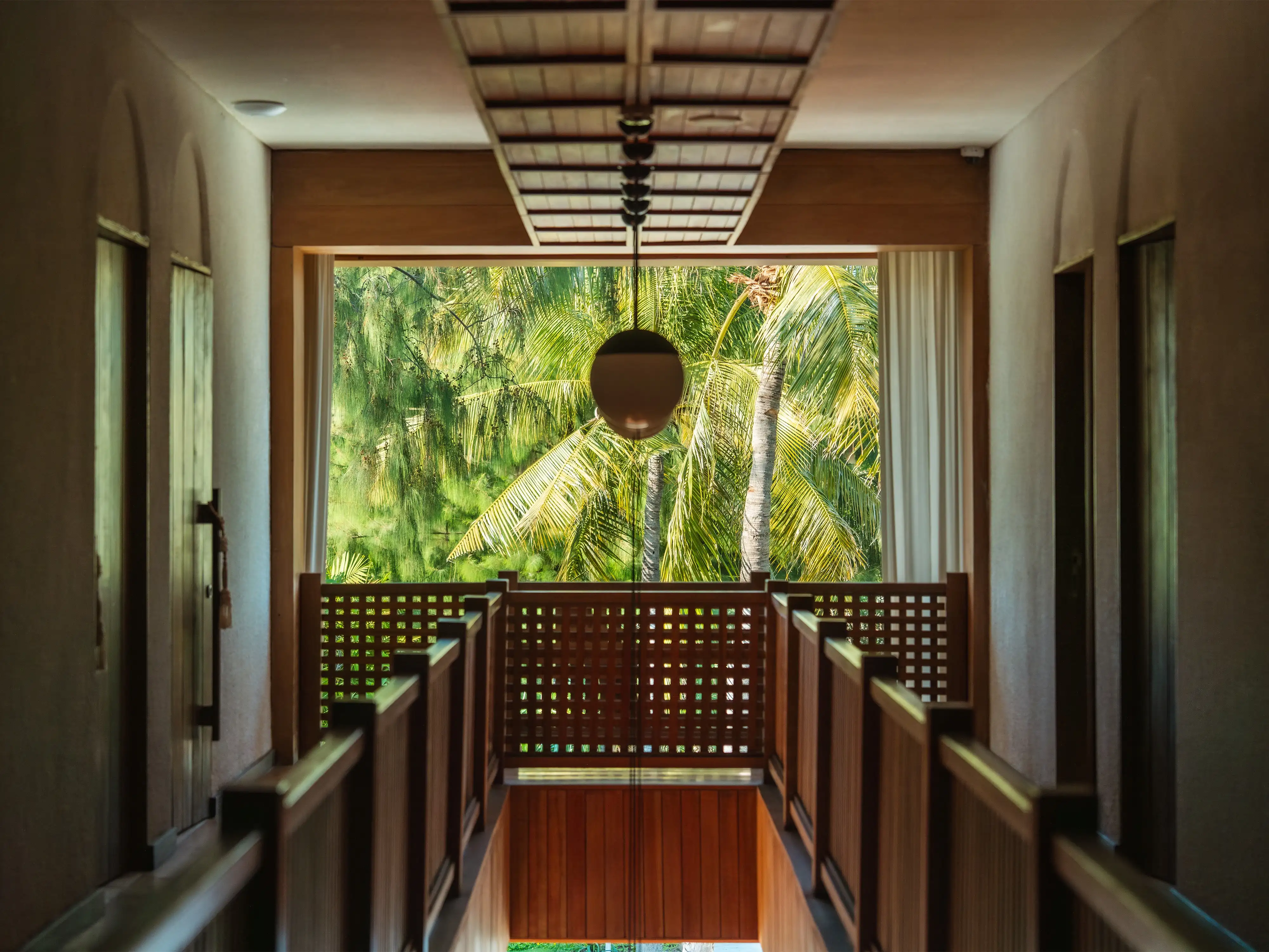 Wooden interior corridor featuring a round pendant light, lattice wooden railings, and a view of lush palm trees at the end of the hall.