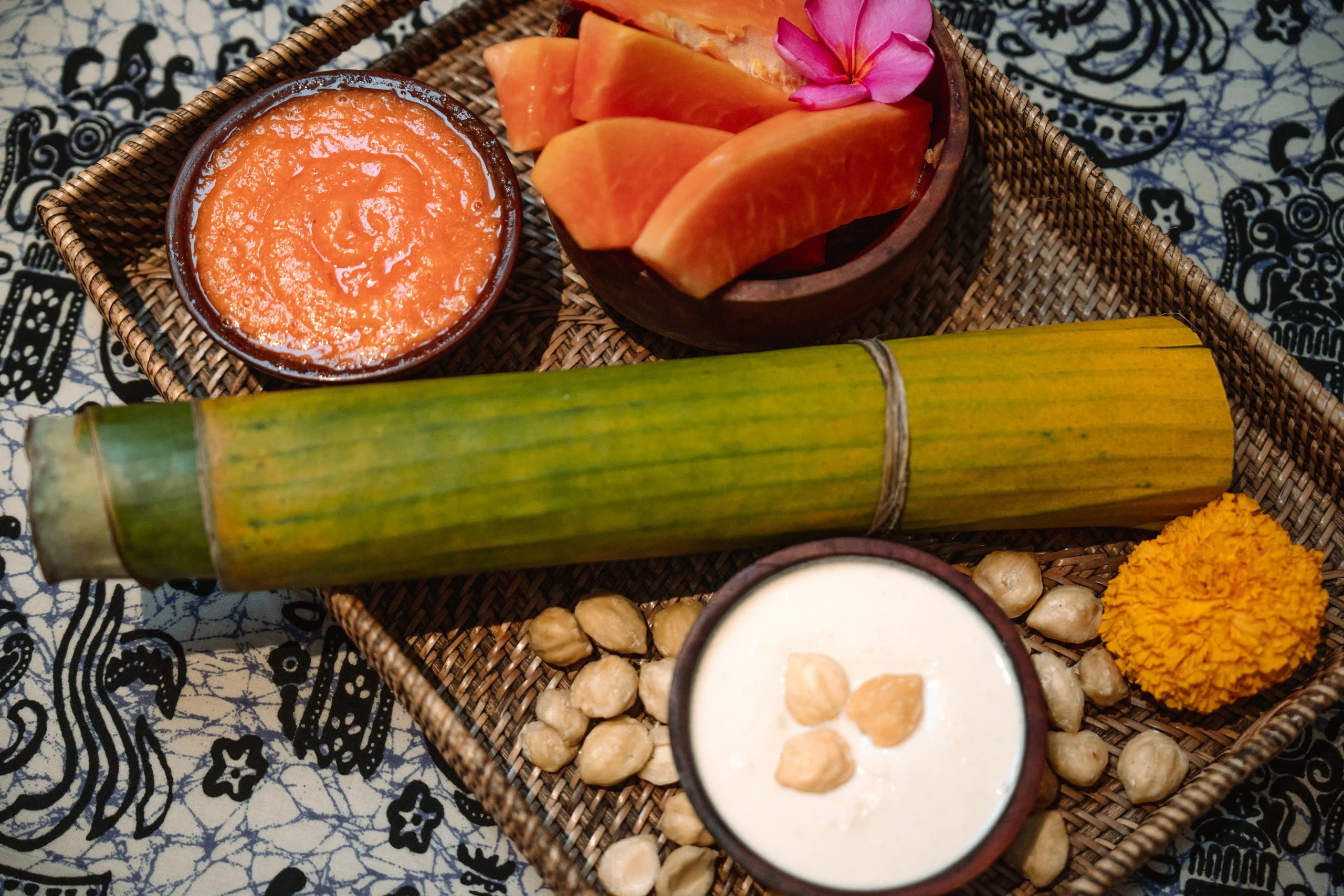 : Flat-lay view of a woven tray containing natural spa ingredients, including papaya slices, scrub paste, candlenuts, a rolled banana leaf, and fresh flowers.