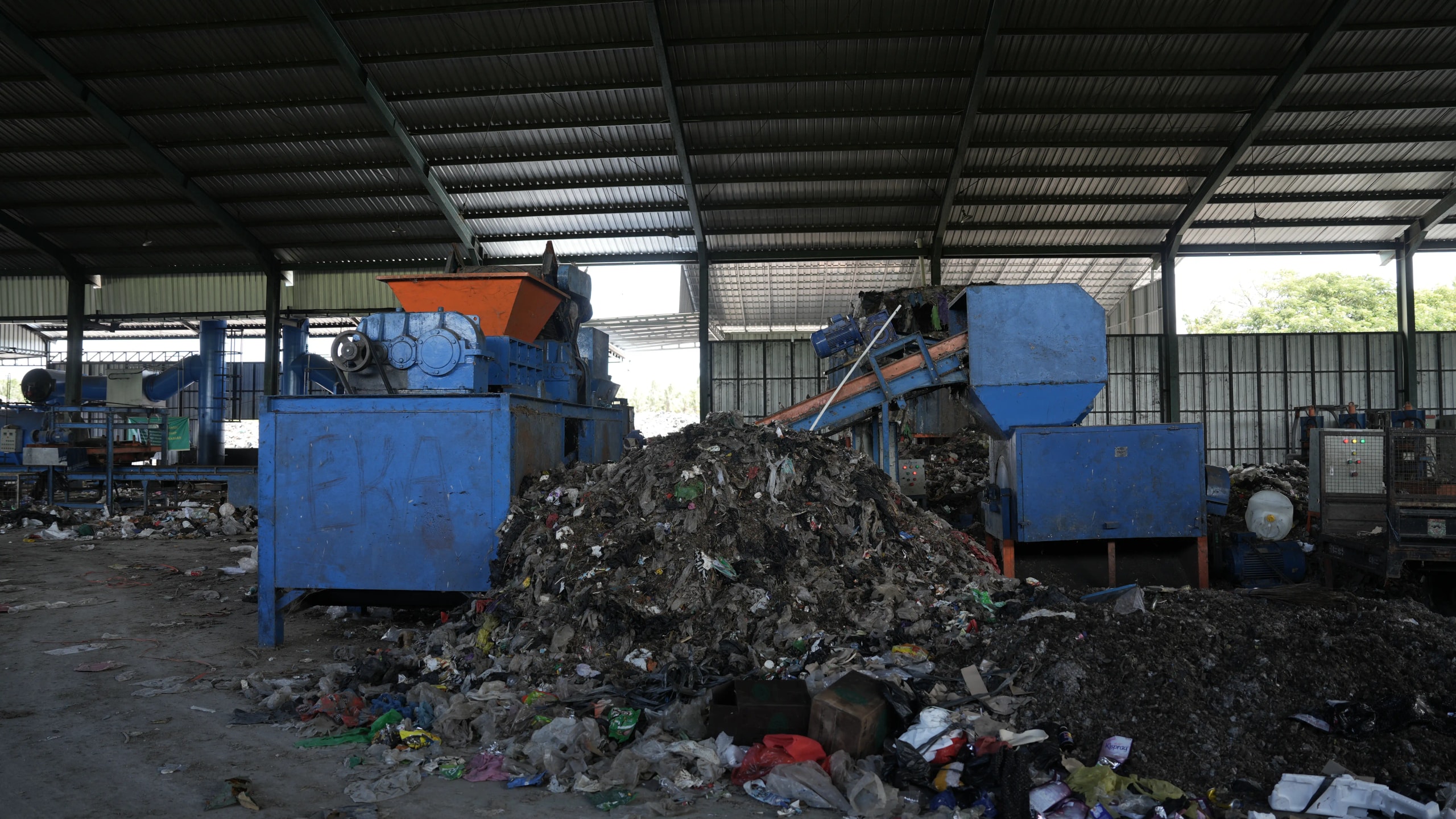 Large blue industrial waste-sorting machinery and conveyor belts next to a massive pile of un-sorted plastic trash inside a high-ceiling facility.