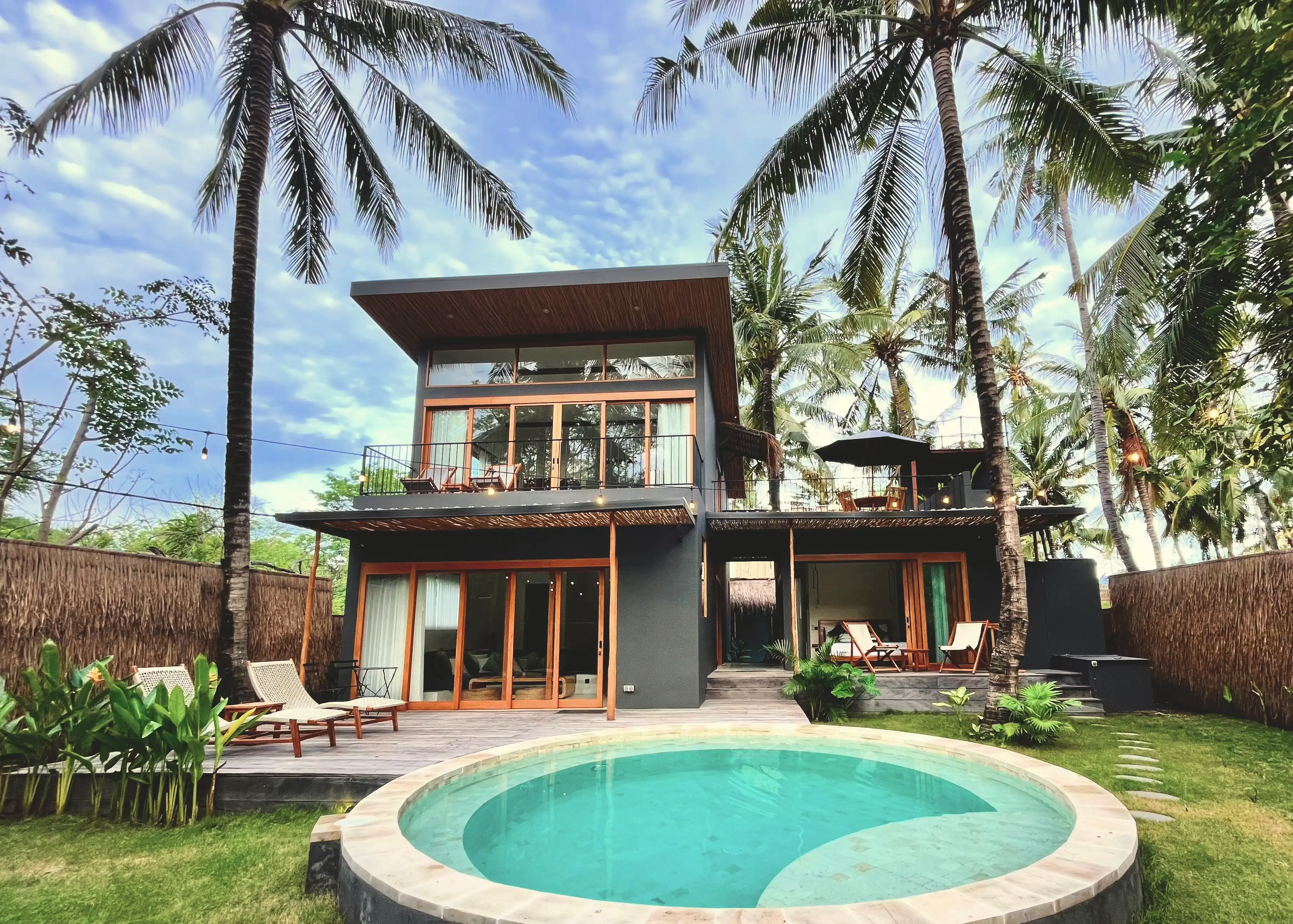 Modern two-story villa exterior featuring a circular plunge pool, wooden patio, and towering palm trees.