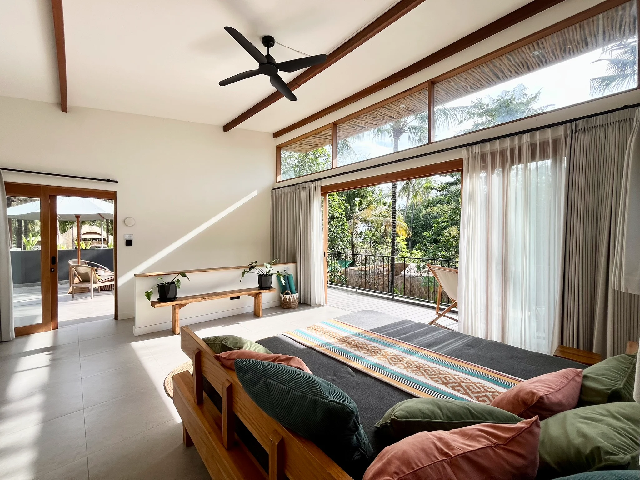 Spacious bedroom with high ceilings, plush daybed seating, wooden beams, and floor-to-ceiling glass doors opening onto a sunny balcony.