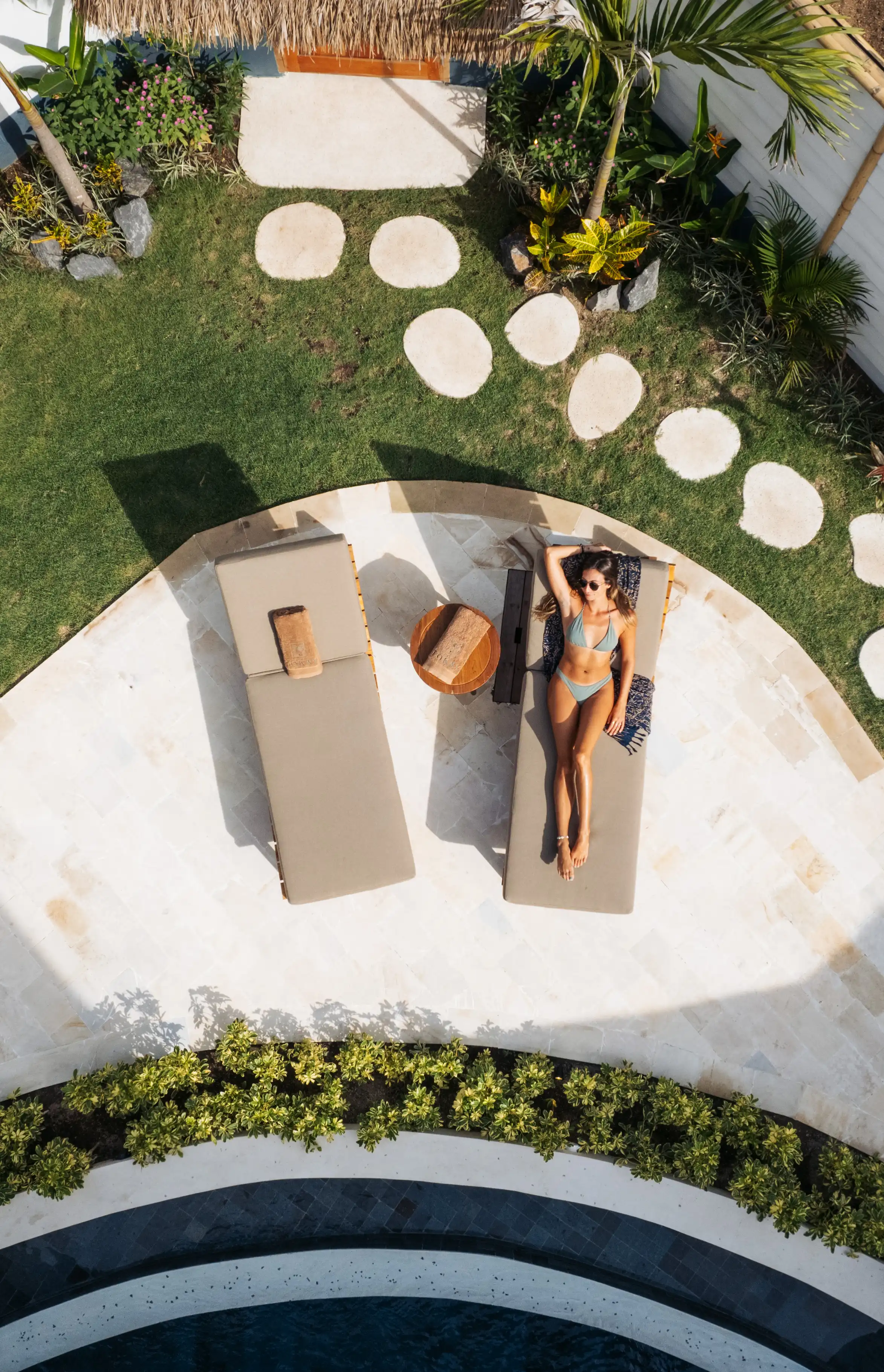 Top-down view of a woman relaxing on a sunbed beside a stone patio and garden pathway.