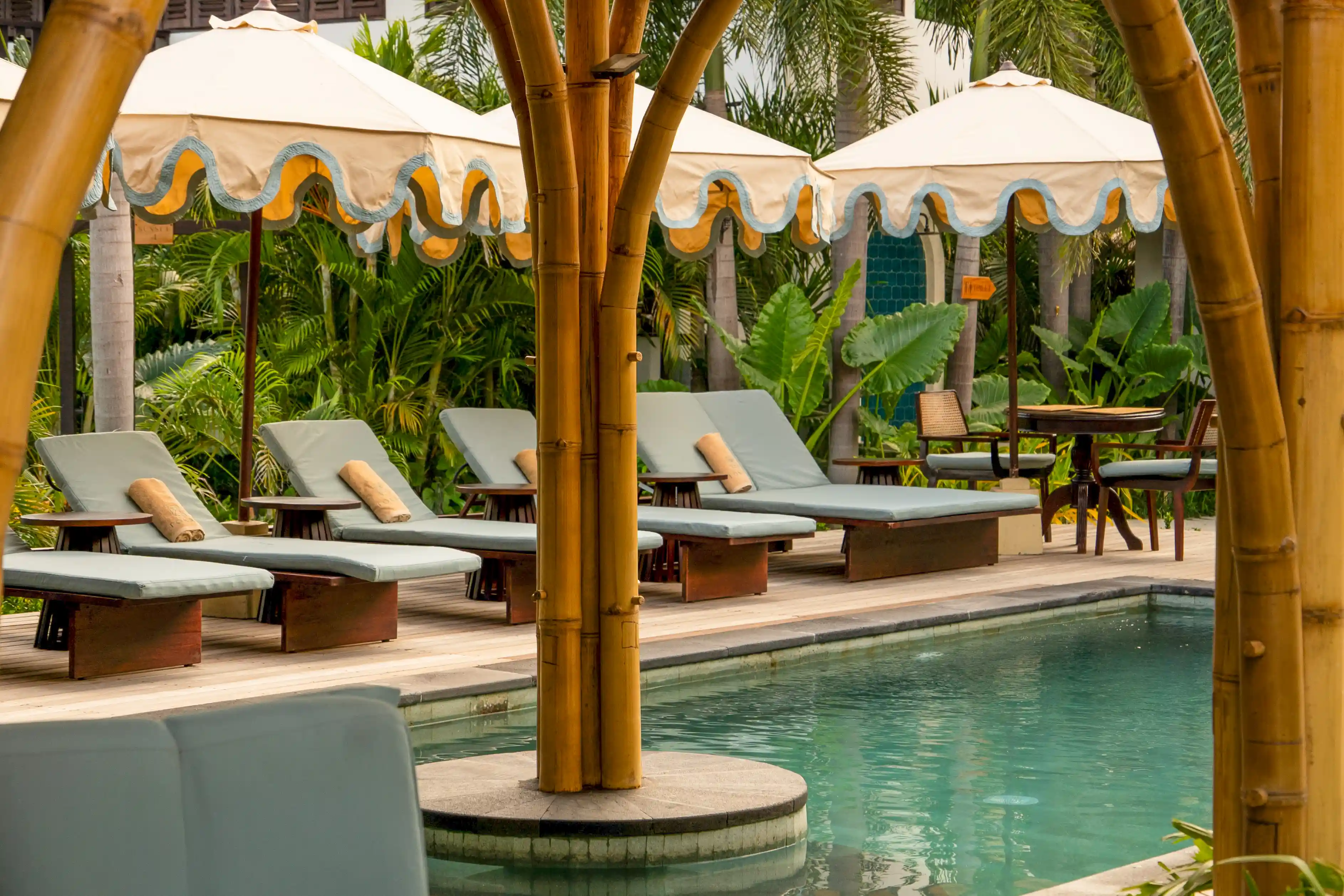 Poolside deck lined with cushioned sun loungers, vintage-style umbrellas, and bamboo pillars.