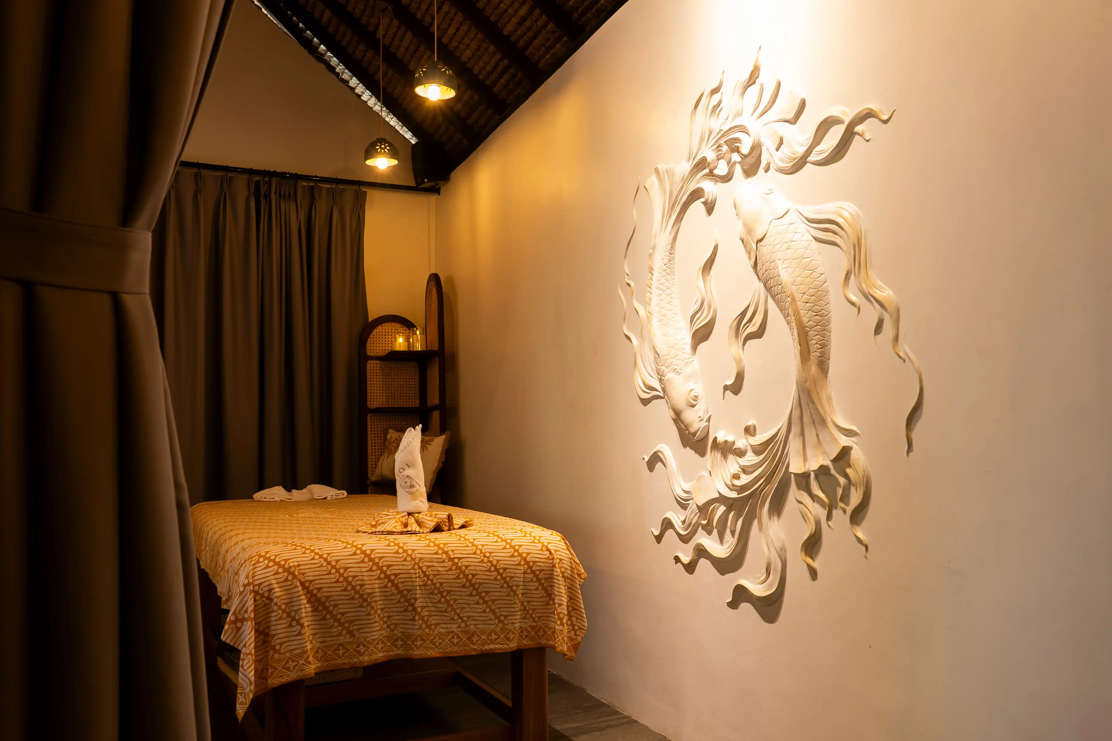 Spa treatment room with a bed dressed in batik fabric and a large carved fish relief on the wall.