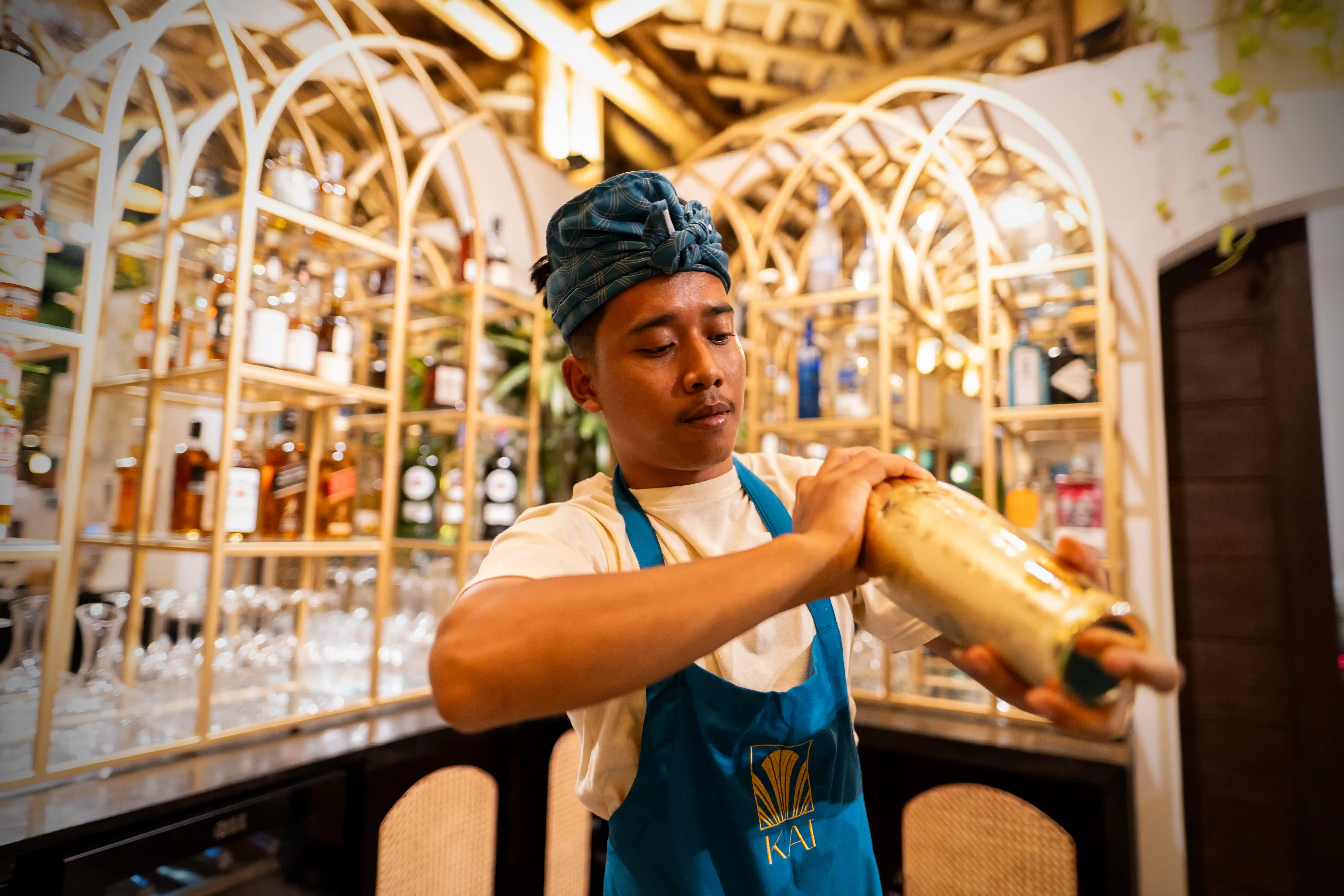 Bartender wearing a traditional headband actively shaking a cocktail in front of an illuminated wooden bar shelf.