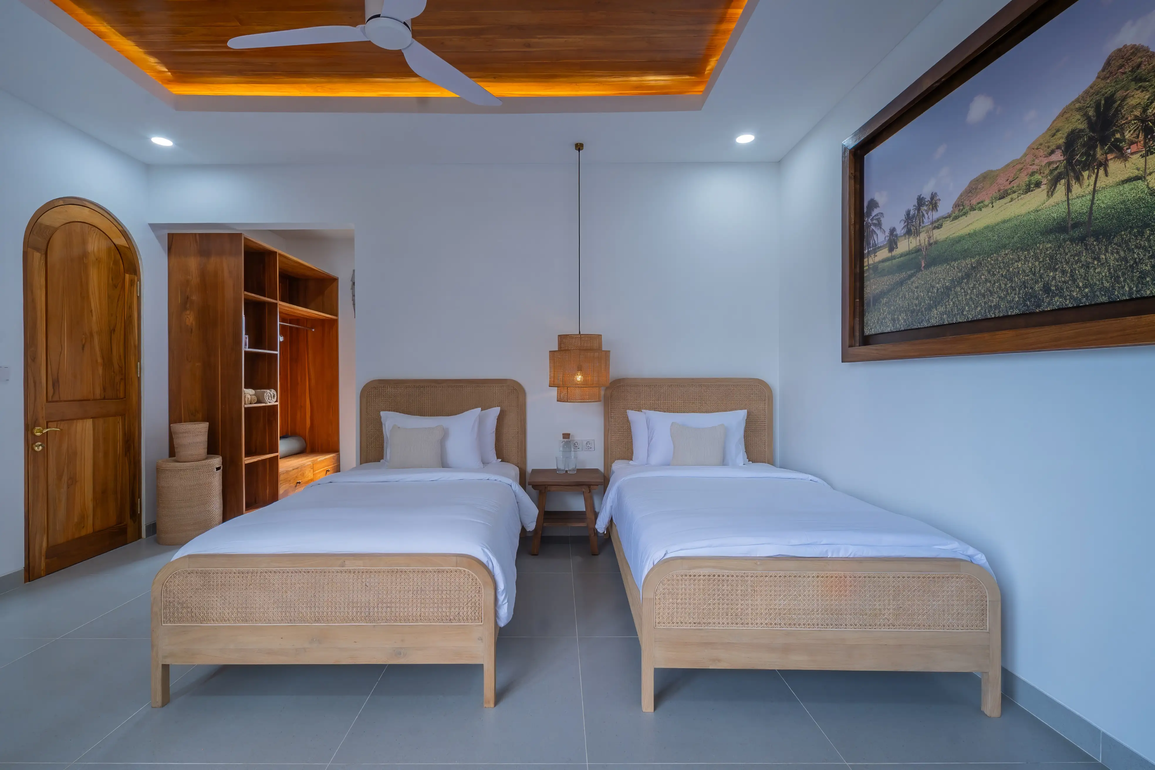 Chambre d'amis avec deux lits simples en rotin, penderie ouverte en bois et grand tableau mural.