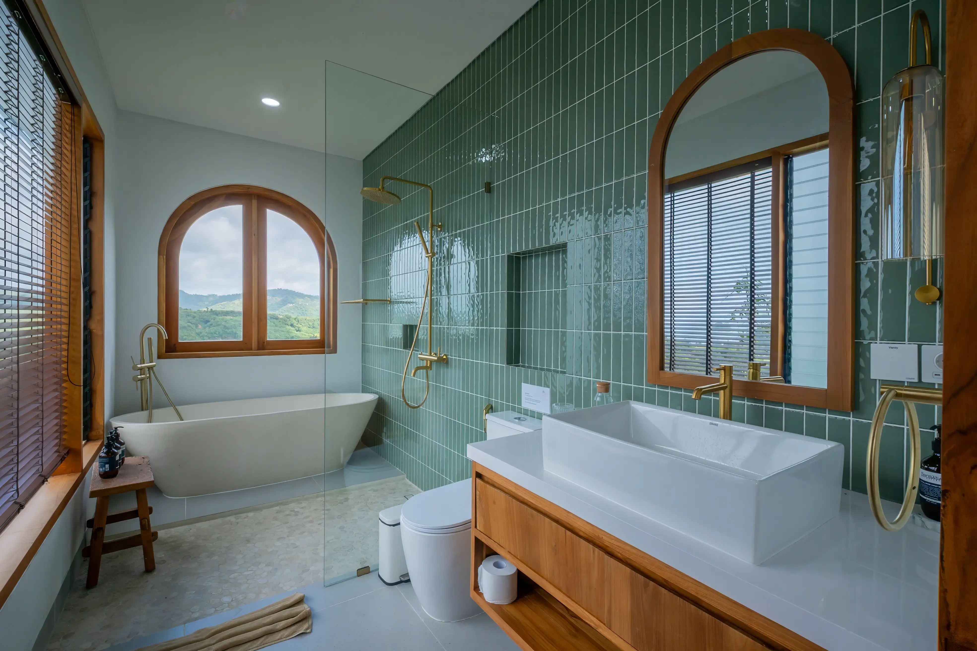 Salle de bain élégante avec carrelage vert, baignoire îlot devant une fenêtre arquée et meuble vasque en bois.