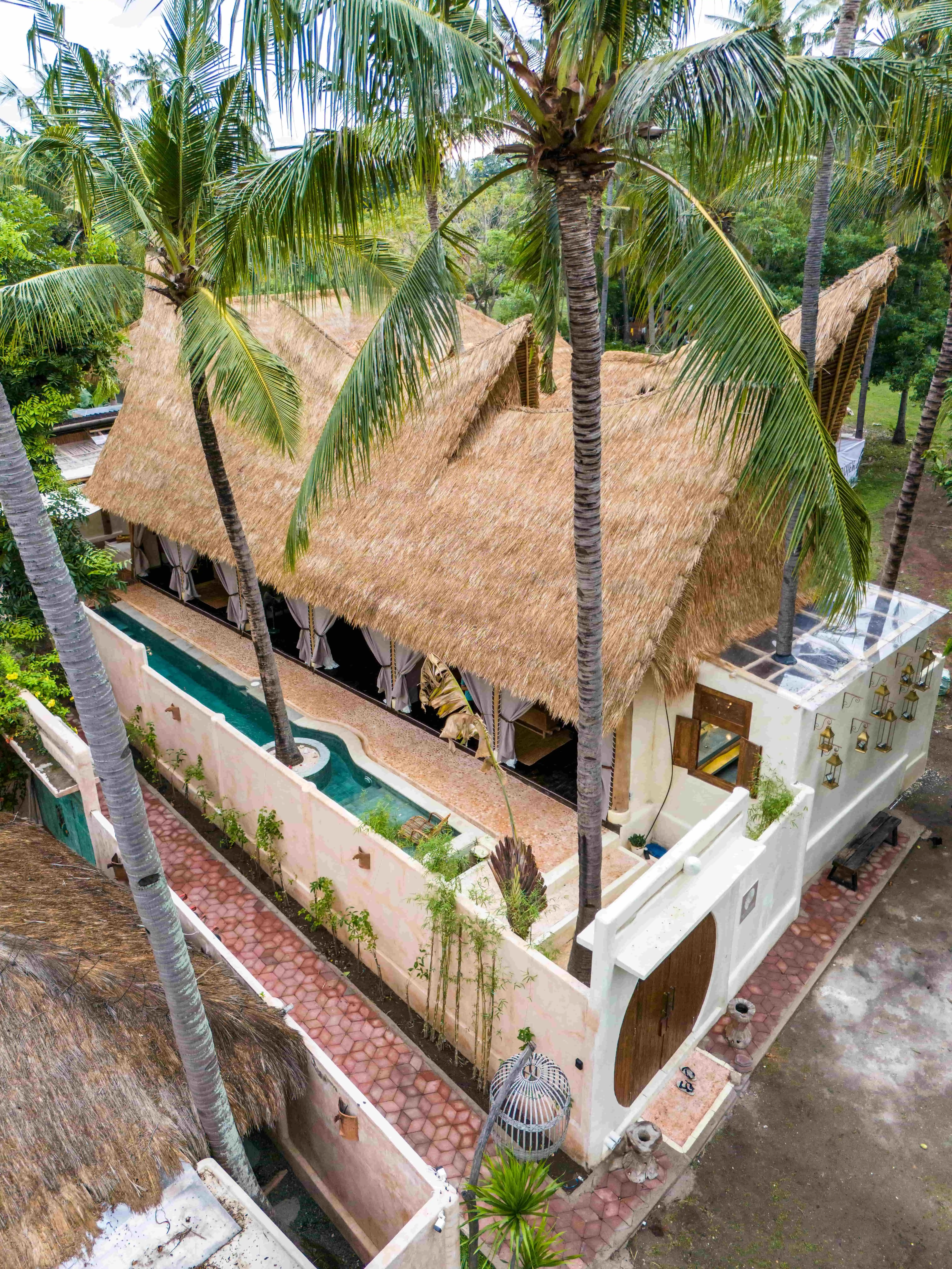 Aerial view of a luxury tropical villa with thatched roofs, a private swimming pool, and lush palm trees.