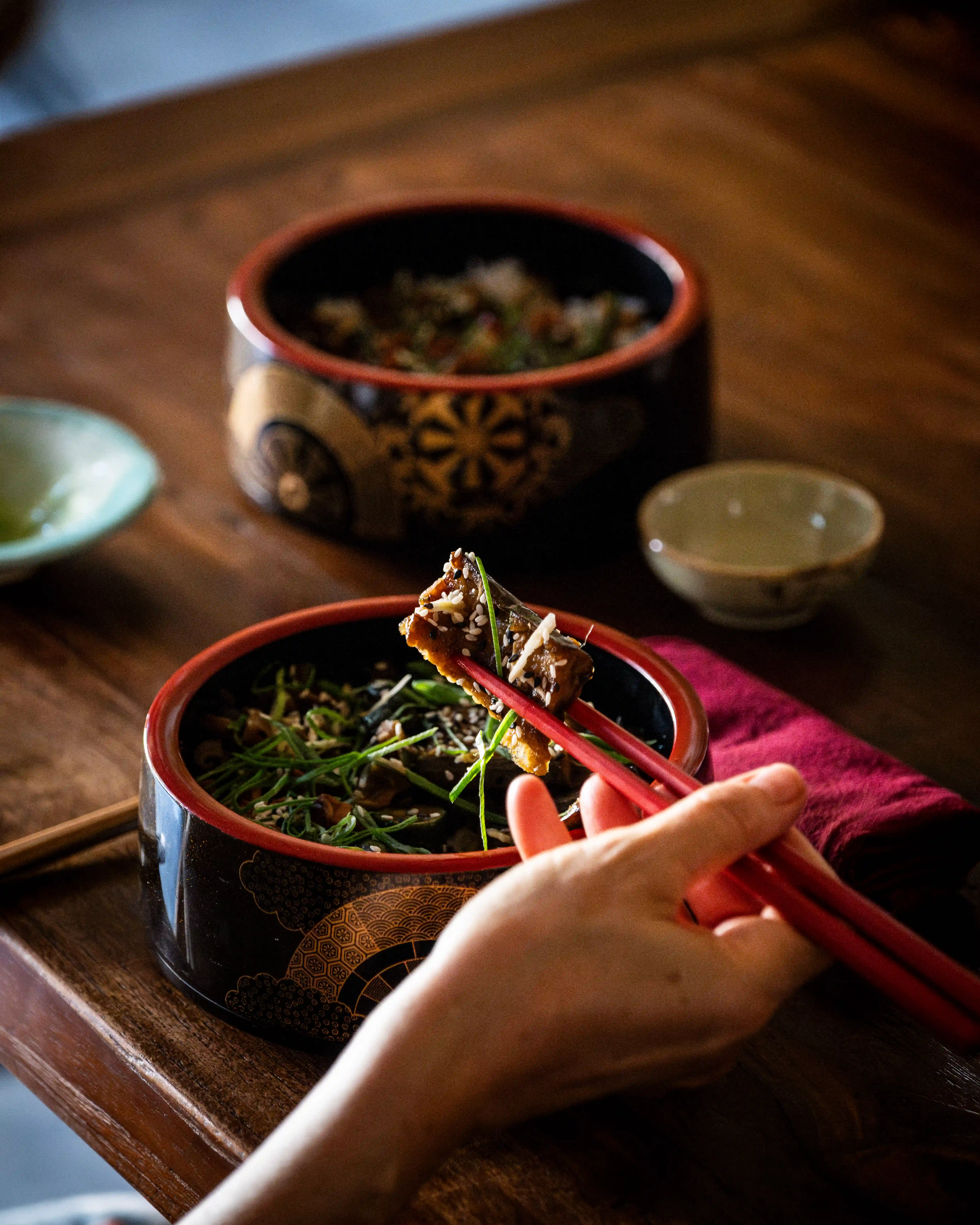 Hand holding red chopsticks taking a piece of grilled eel from a lacquer bento box.