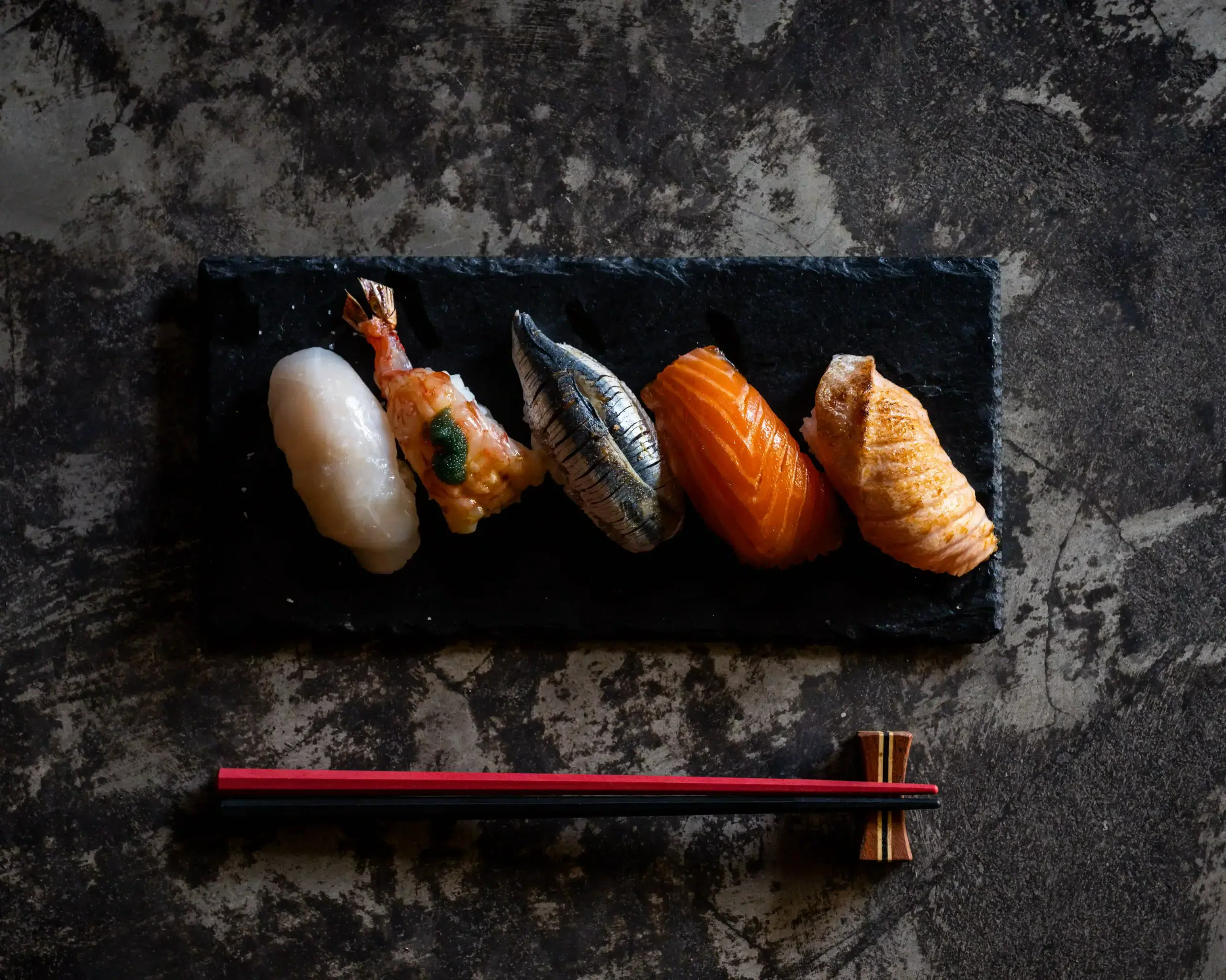 Five pieces of assorted nigiri sushi arranged vertically on a black slate platter.
