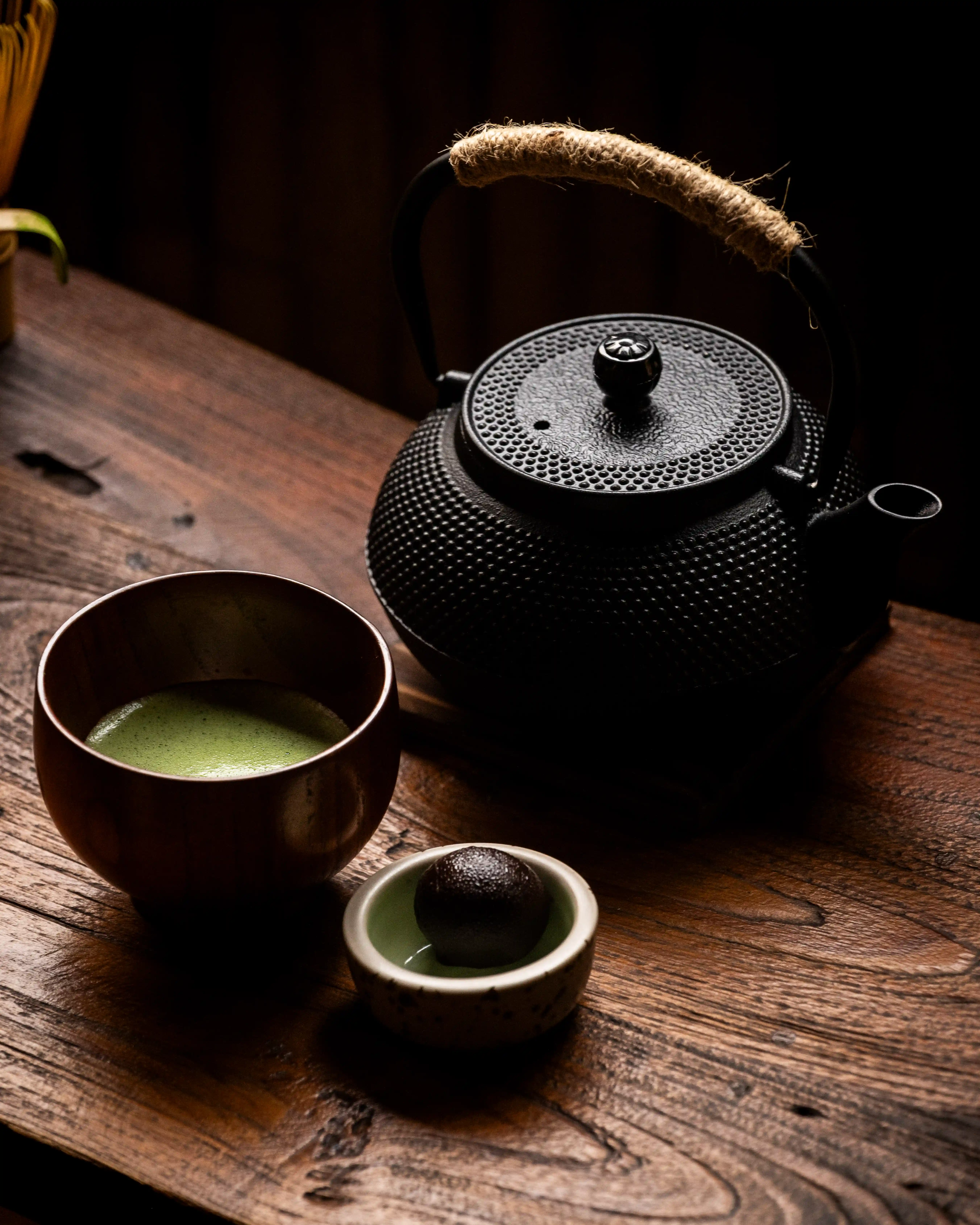 A black cast-iron teapot, a wooden cup of matcha green tea, and a sweet treat on a wooden table.