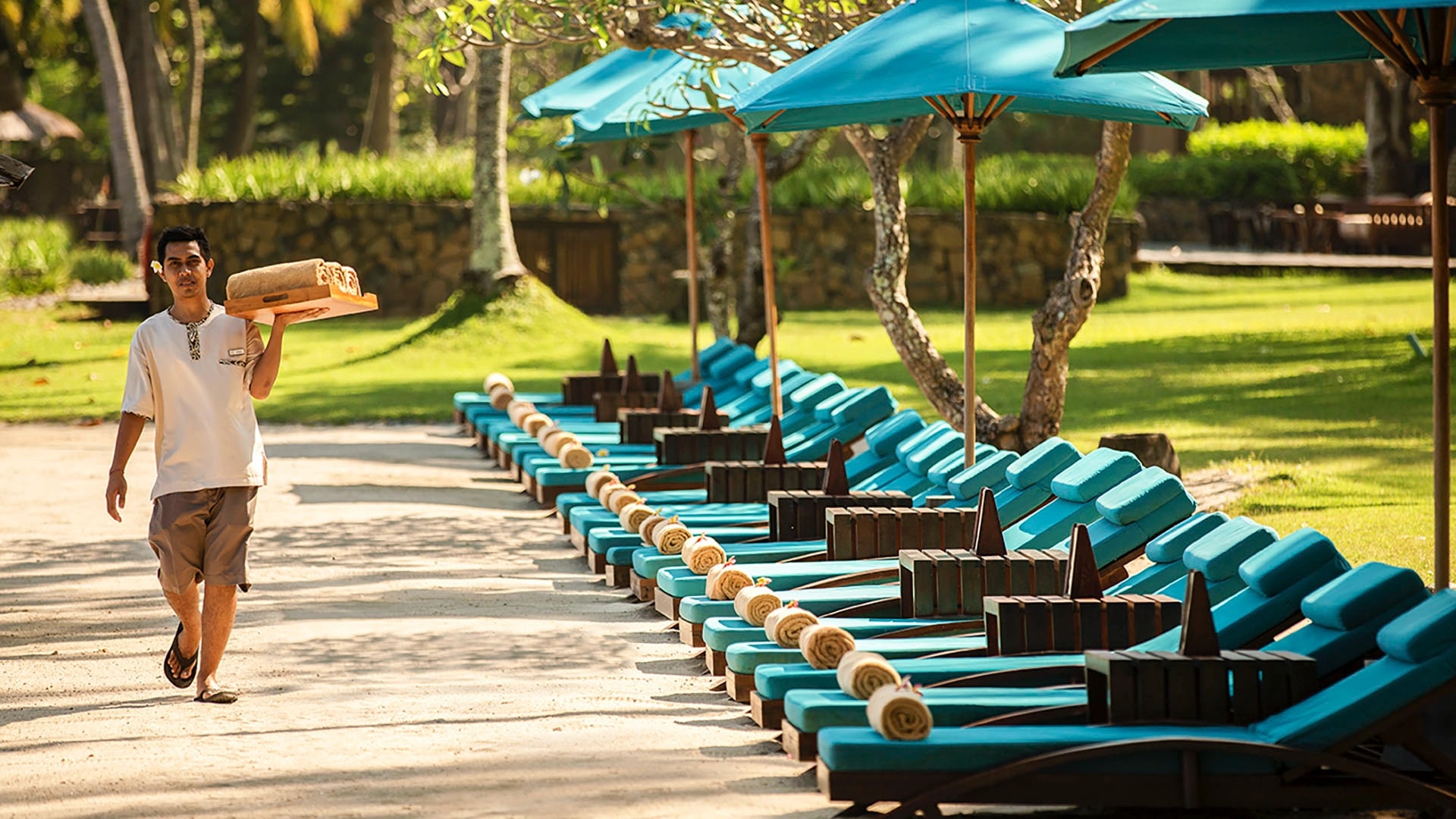 Resort staff member carrying rolled towels along a row of blue sun loungers under turquoise umbrellas on the beach.