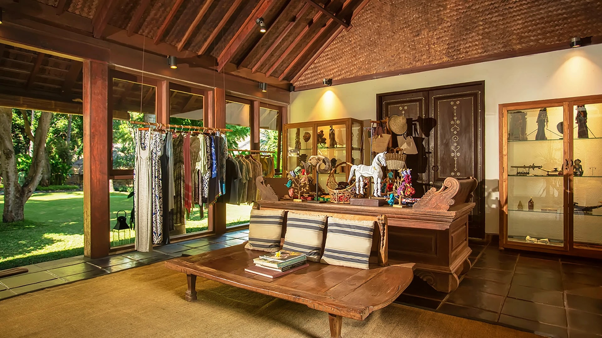 Resort boutique shop displaying clothing racks, local handicrafts, wooden statues, and glass display cases under a thatched roof.