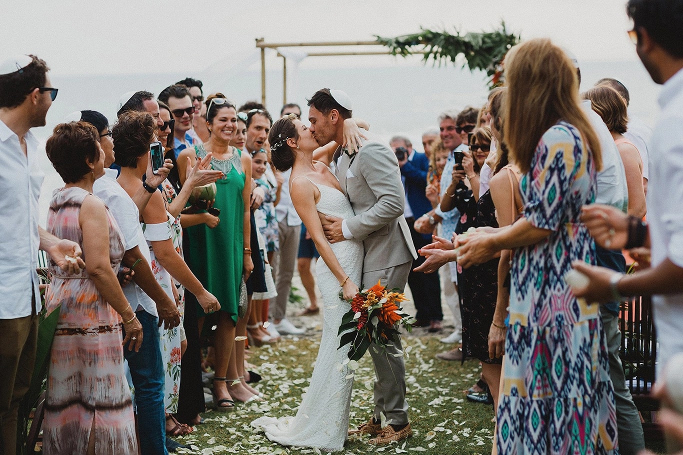 Bride and groom kissing under a beachside wedding arch while guests cheer and throw flower petals.