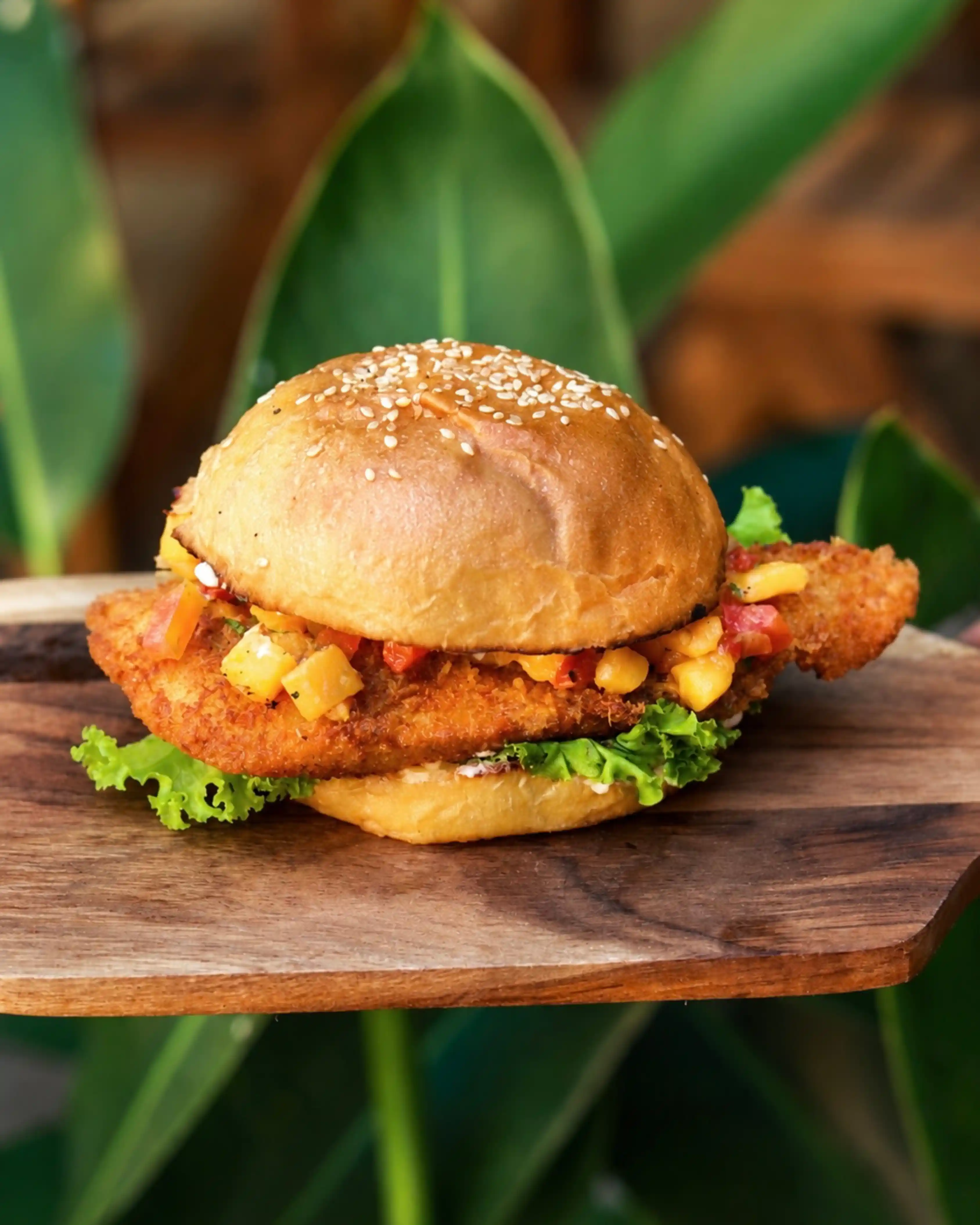 A crispy fried chicken burger served on a wooden board, topped with mango salsa and fresh green lettuce.