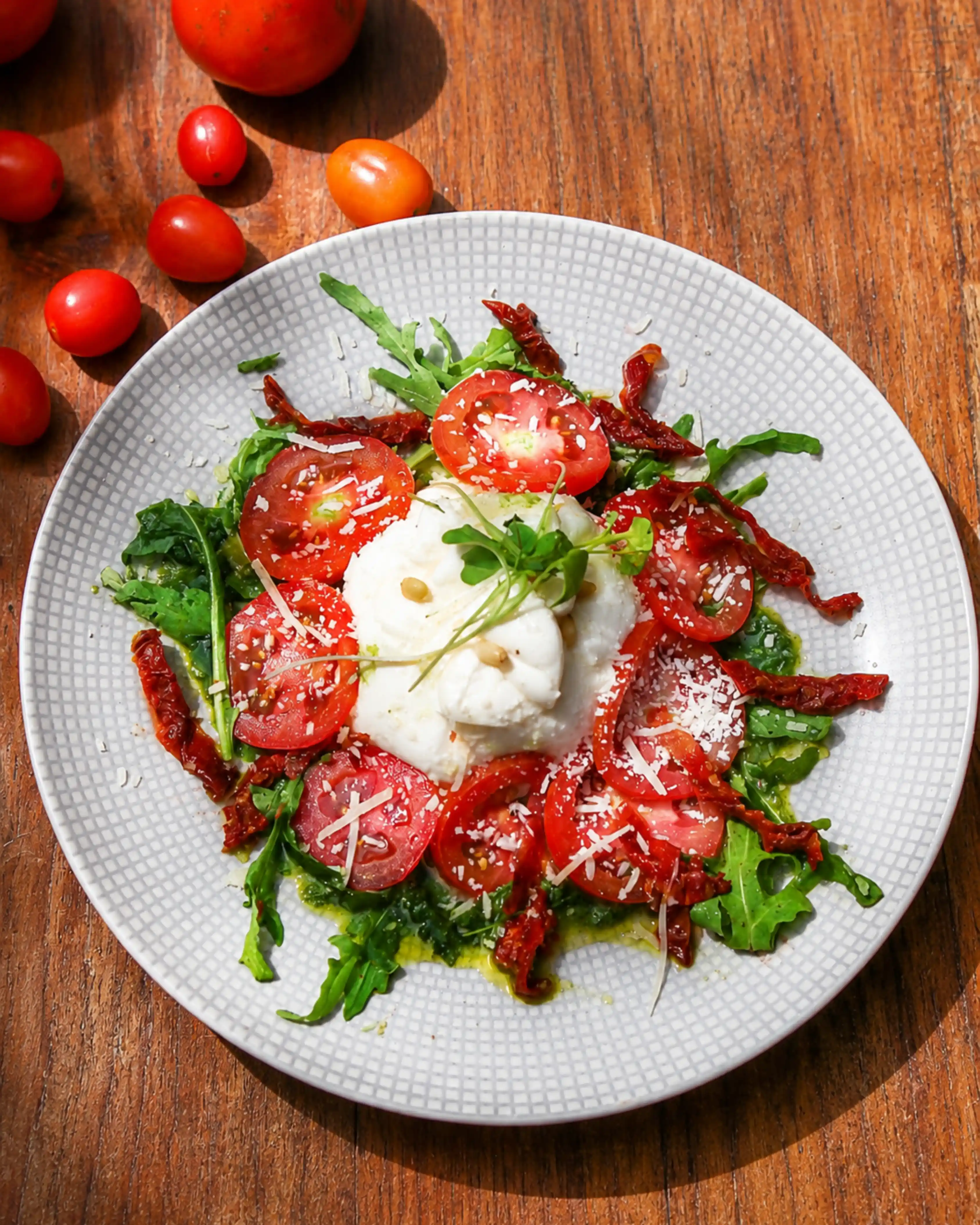 A plated caprese salad featuring fresh burrata cheese in the center, surrounded by sliced fresh tomatoes, sun-dried tomatoes, and arugula leaves.