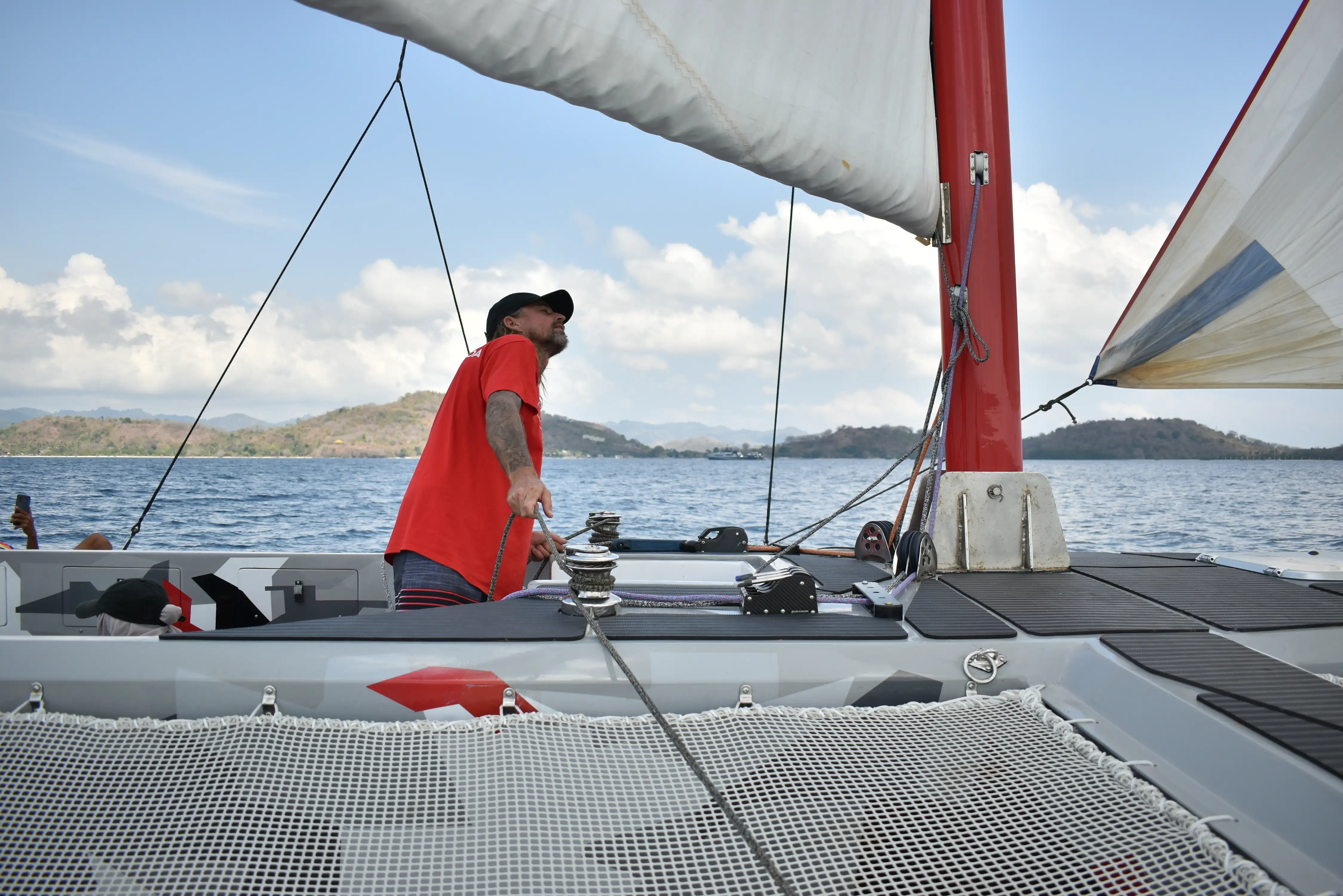 : A sailor adjusting the winch on the deck of a trimaran boat with islands in the background.