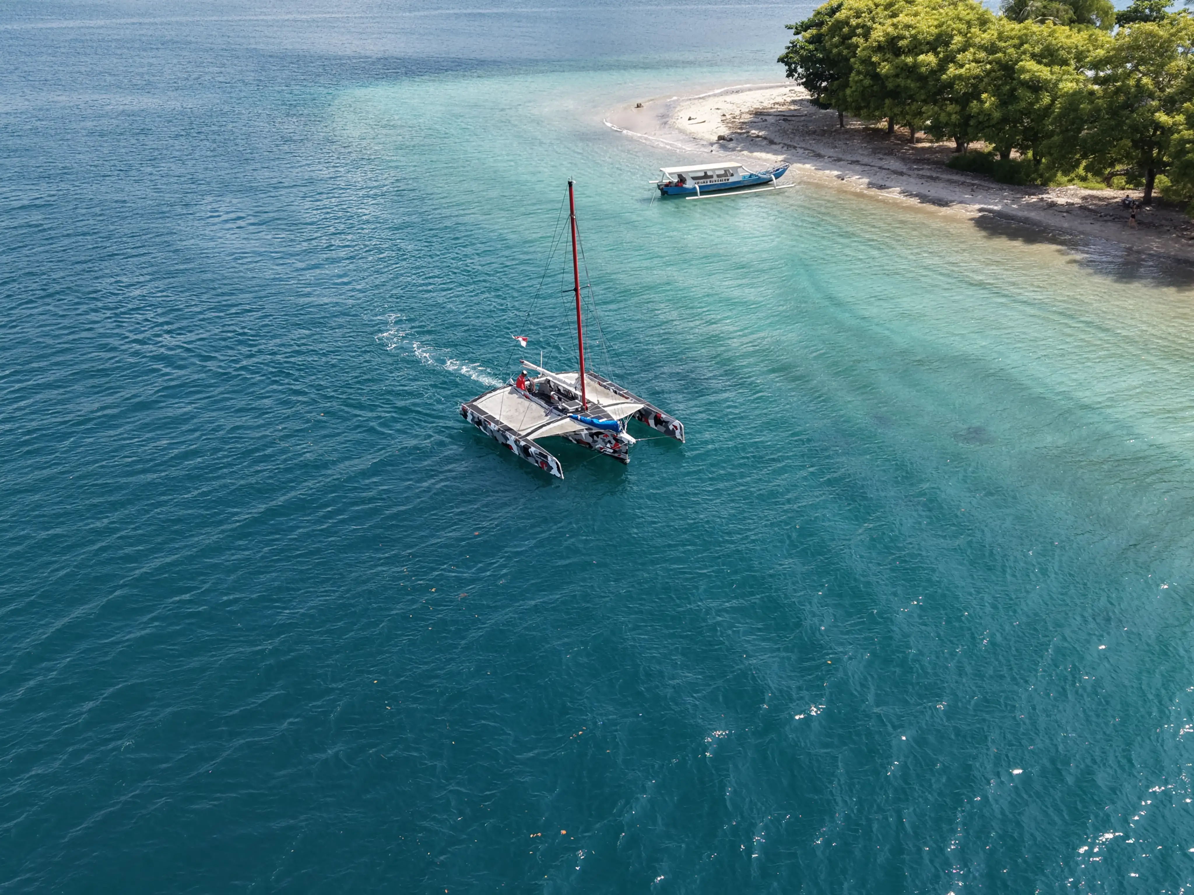 Aerial view of a trimaran anchored near a white sand beach and tropical greenery.