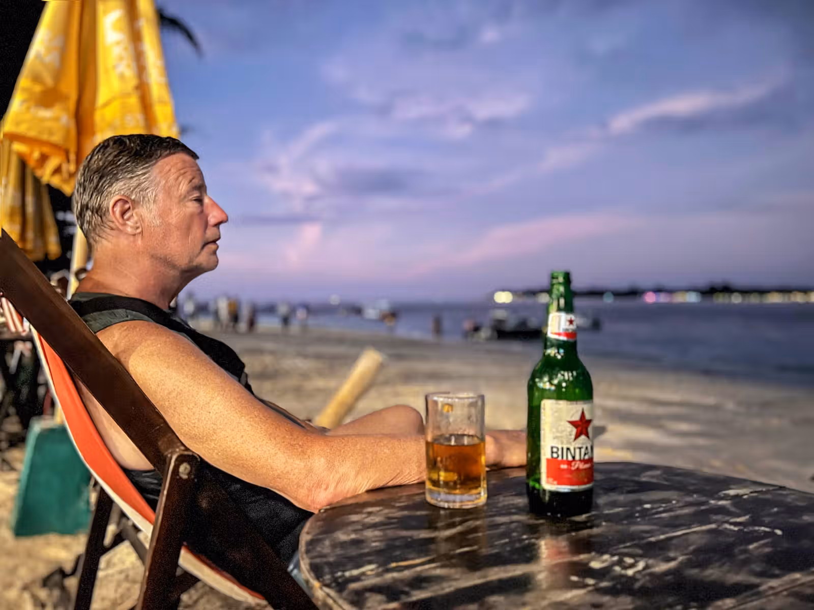 A man relaxing in a beach chair at dusk next to a table with a glass and bottle of beer.