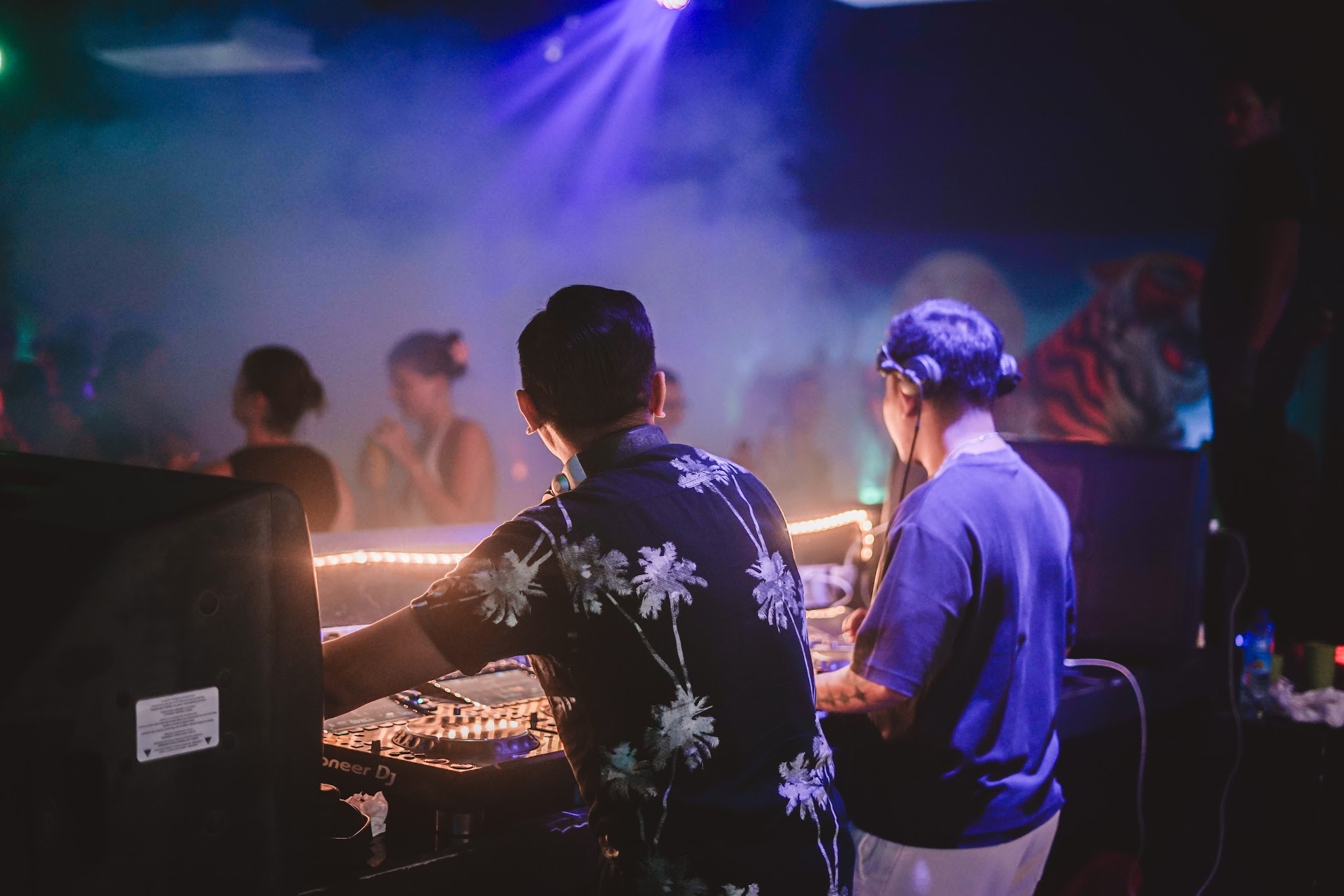 Rear view of two DJs performing at a mixing booth in a dimly lit club filled with blue stage lighting and smoke.