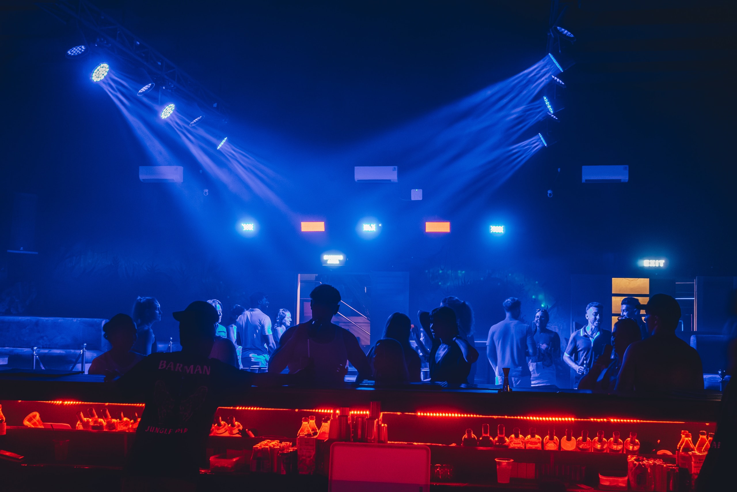 Silhouettes of partygoers gathered around a bar under blue spotlights inside a crowded nightclub.