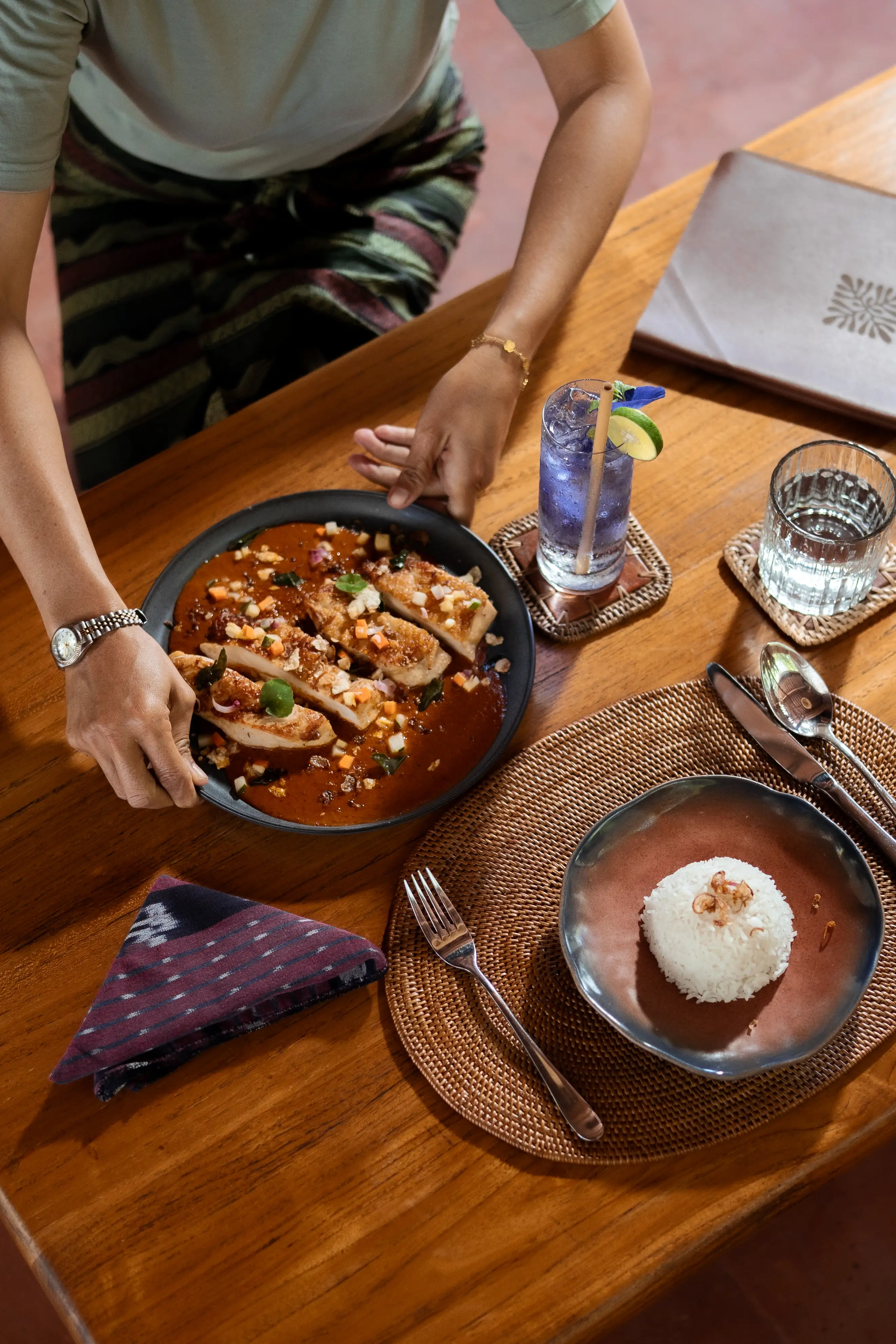 A person placing a plated fish dish with sauce on a wooden table set with rice and drinks.