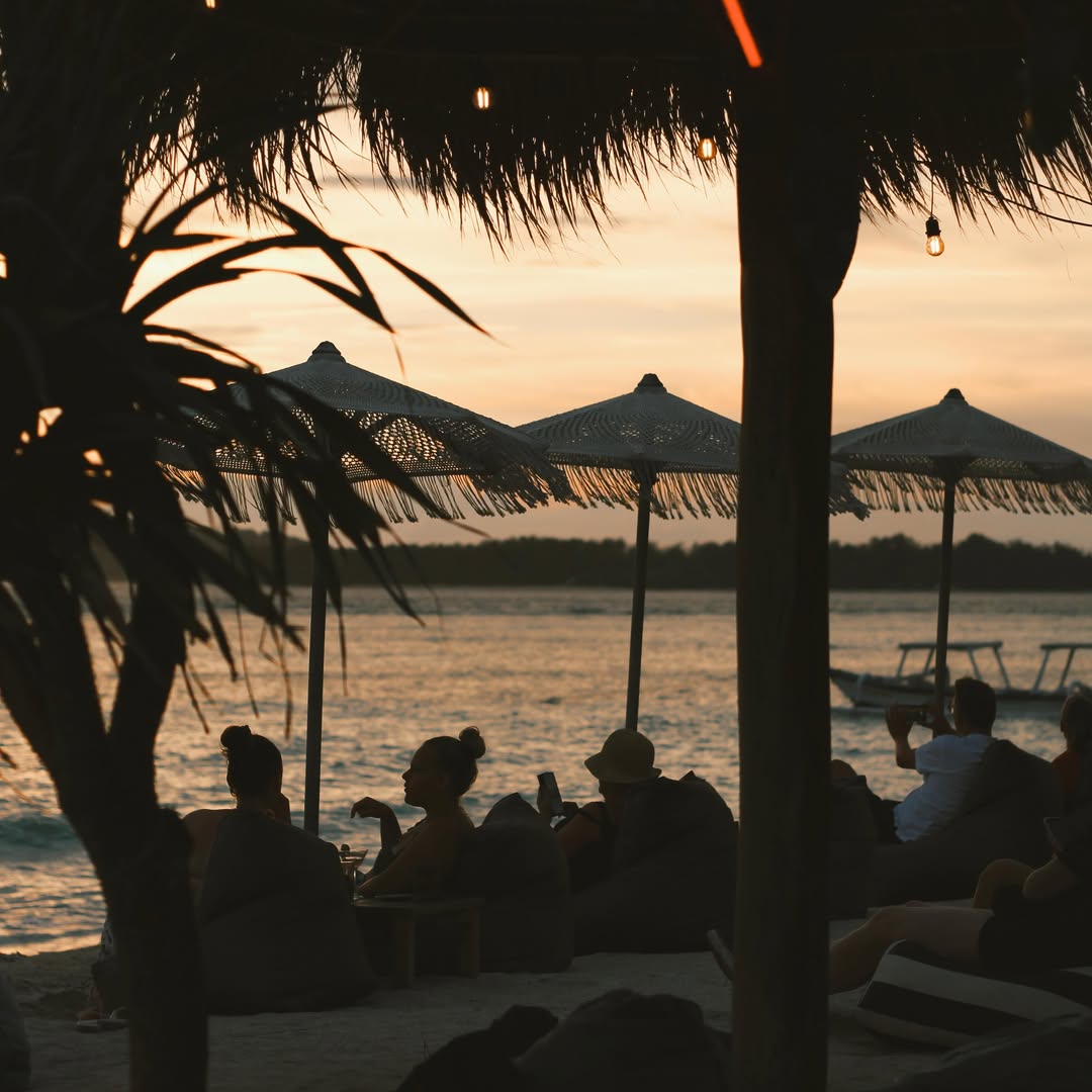 People sitting on beach beanbags under thatched umbrellas during a golden ocean sunset.