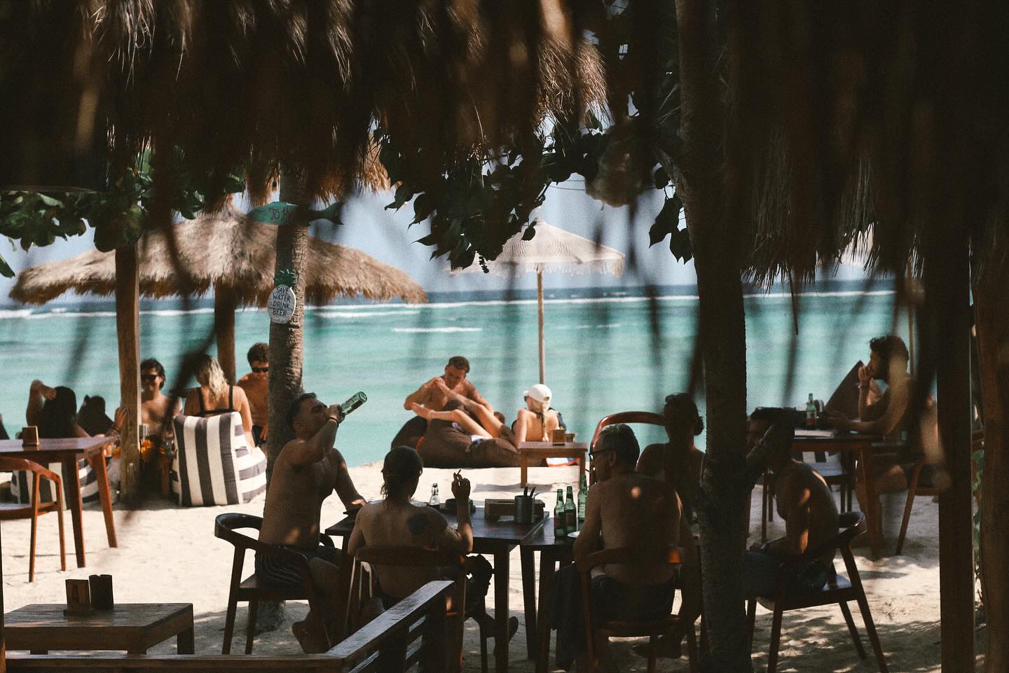 Crowd drinking at wooden beach tables with thatched umbrellas and turquoise water.