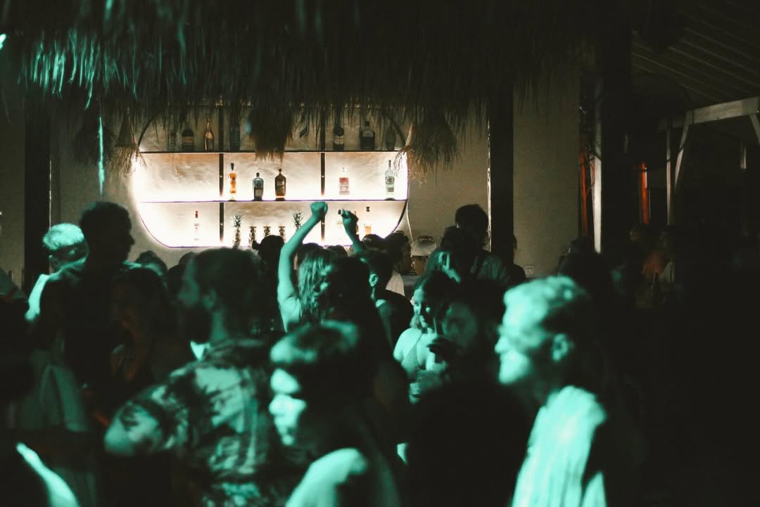 People dancing inside a dimly lit bar with green lights and a glowing bottle shelf.