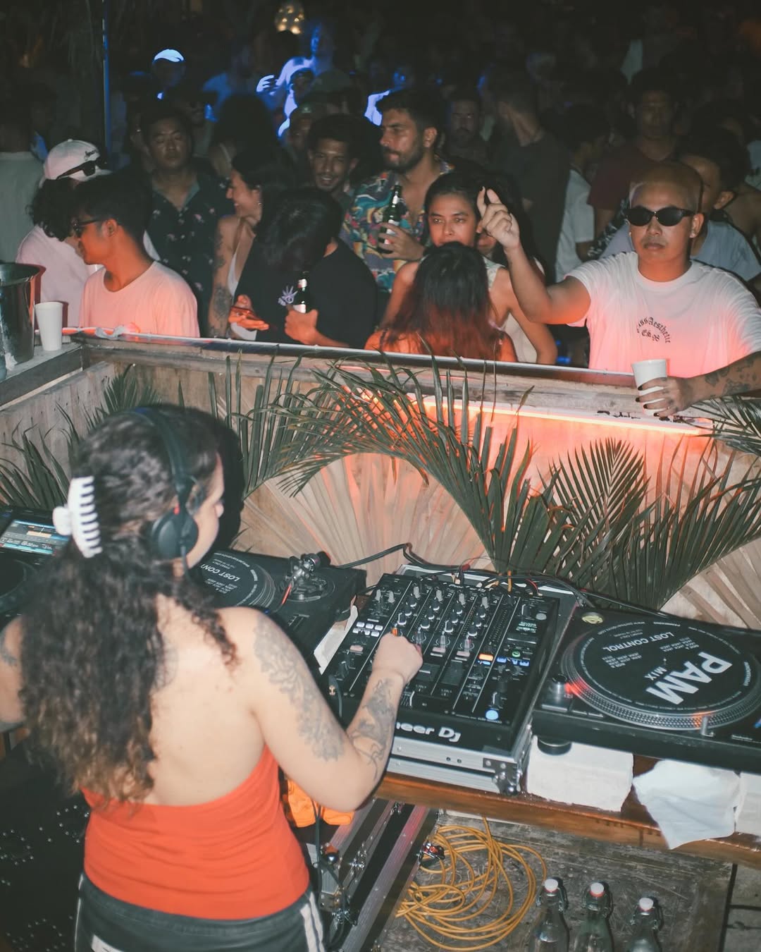 A female DJ at a mixer performing for a crowd under warm party lights.
