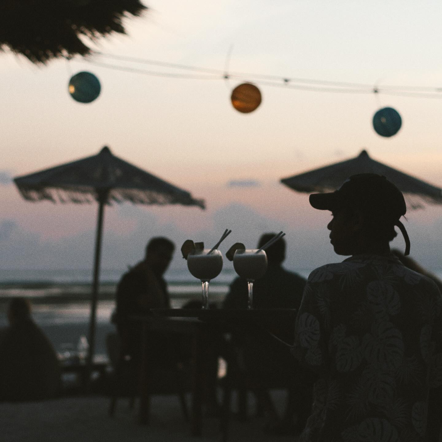 Waiter carrying cocktails on a beach at twilight with silhouetted patrons behind.