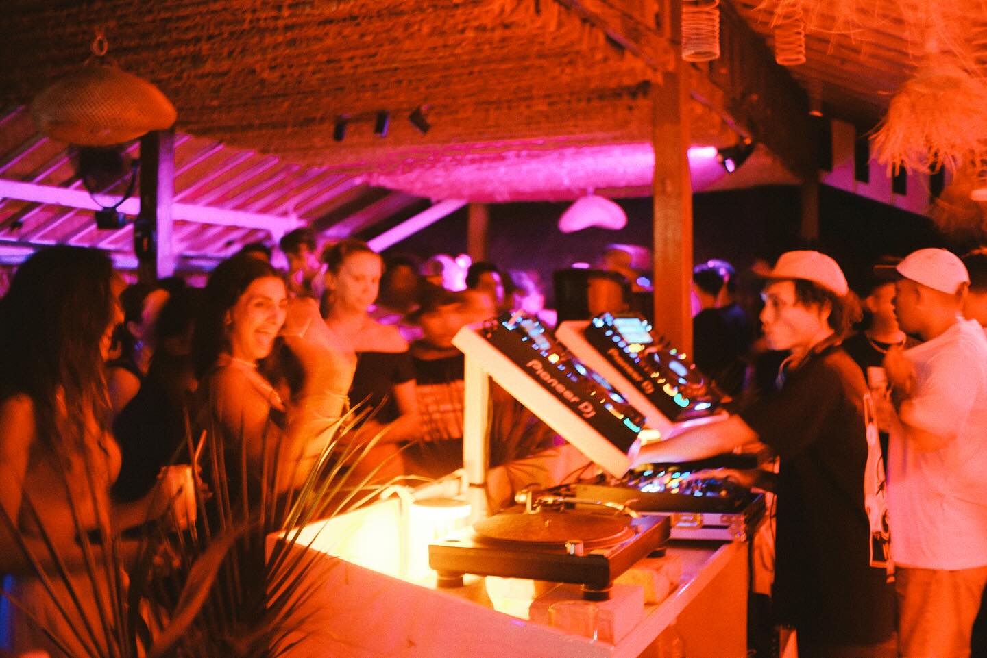 DJ performing at a booth while a crowd dances under orange canopy lights.