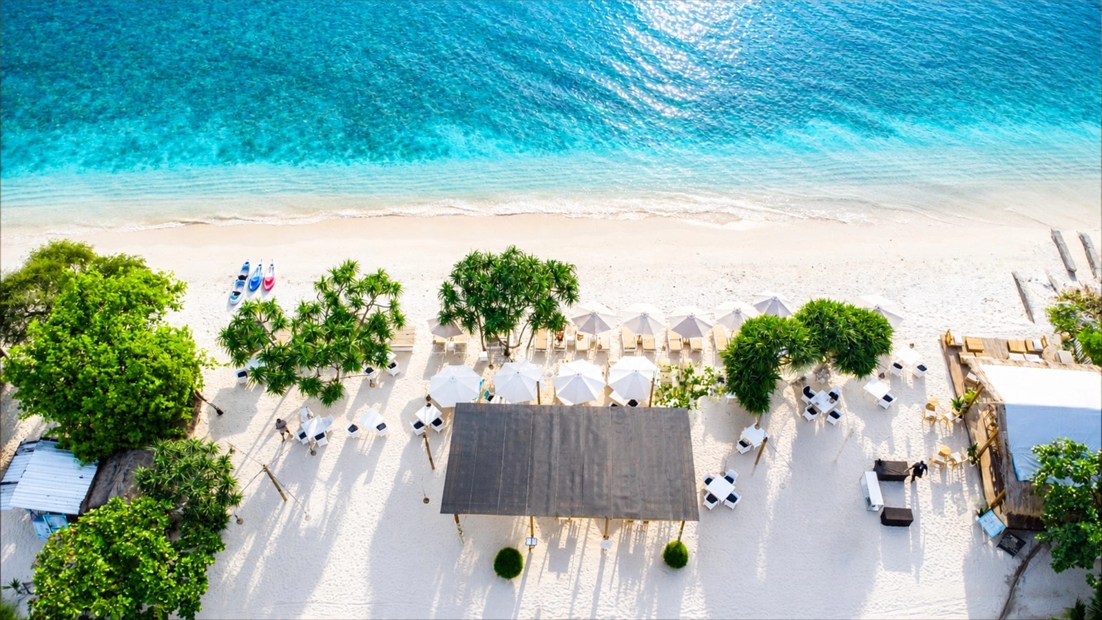 Aerial view of a white sandy beach with umbrellas, palm trees, and turquoise water.