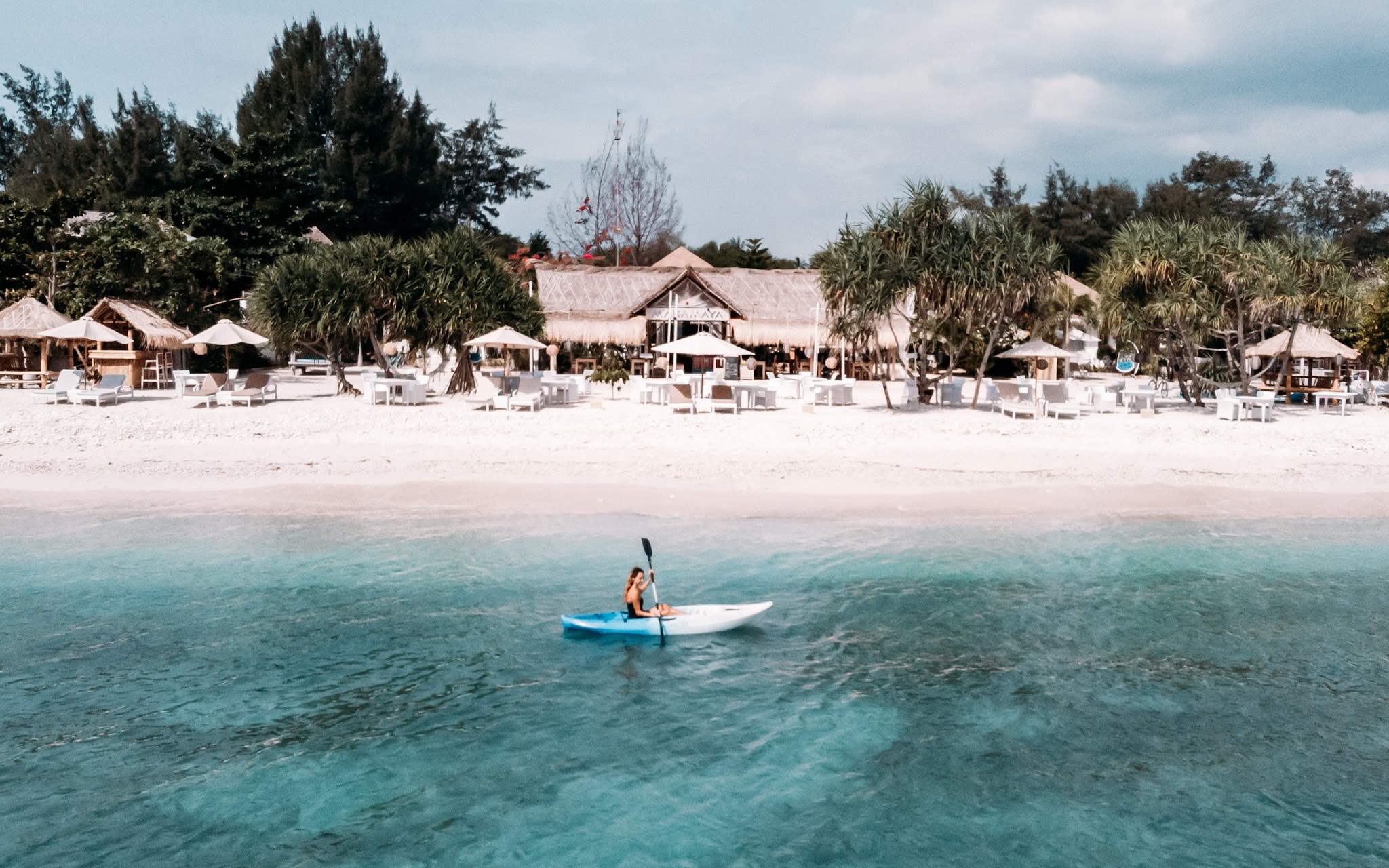 A woman kayaking in clear turquoise ocean water along a beachfront resort.