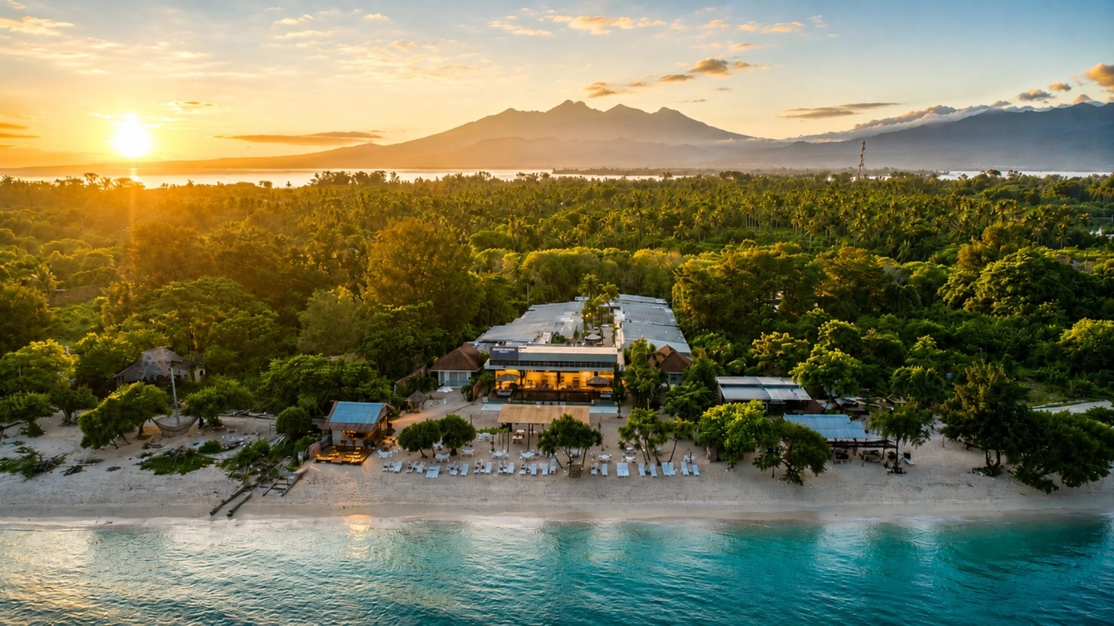 Aerial sunset view of a beachfront resort surrounded by lush trees and mountains.