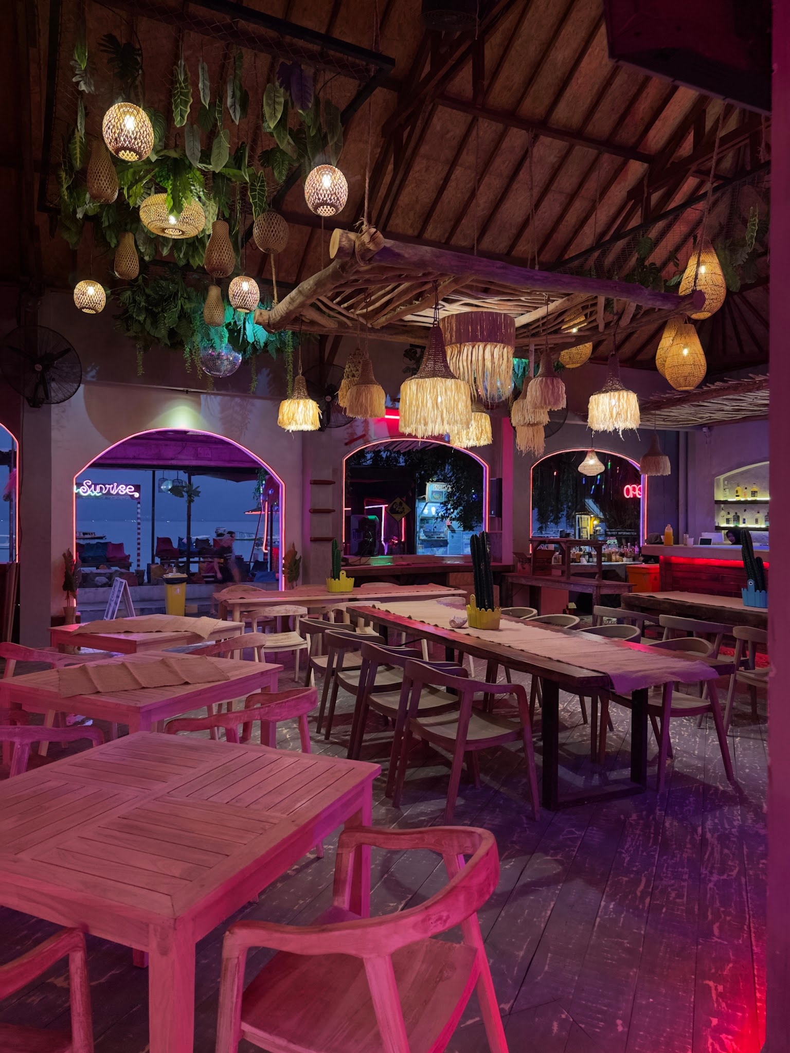 Warm, pink-lit restaurant interior with rustic wooden tables and hanging straw lamps.