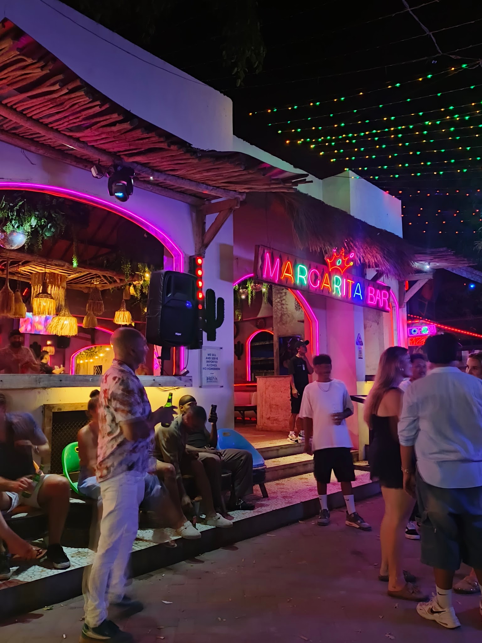 People gathered outside a thatched-roof bar at night under a bright "MARGARITA BAR" neon sign.