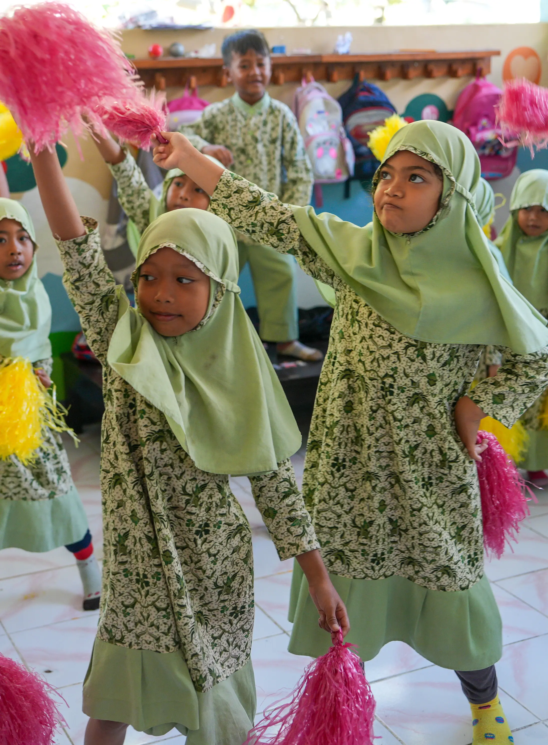 Young schoolgirls in green printed uniforms dancing enthusiastically with bright pink pom-poms inside a classroom.