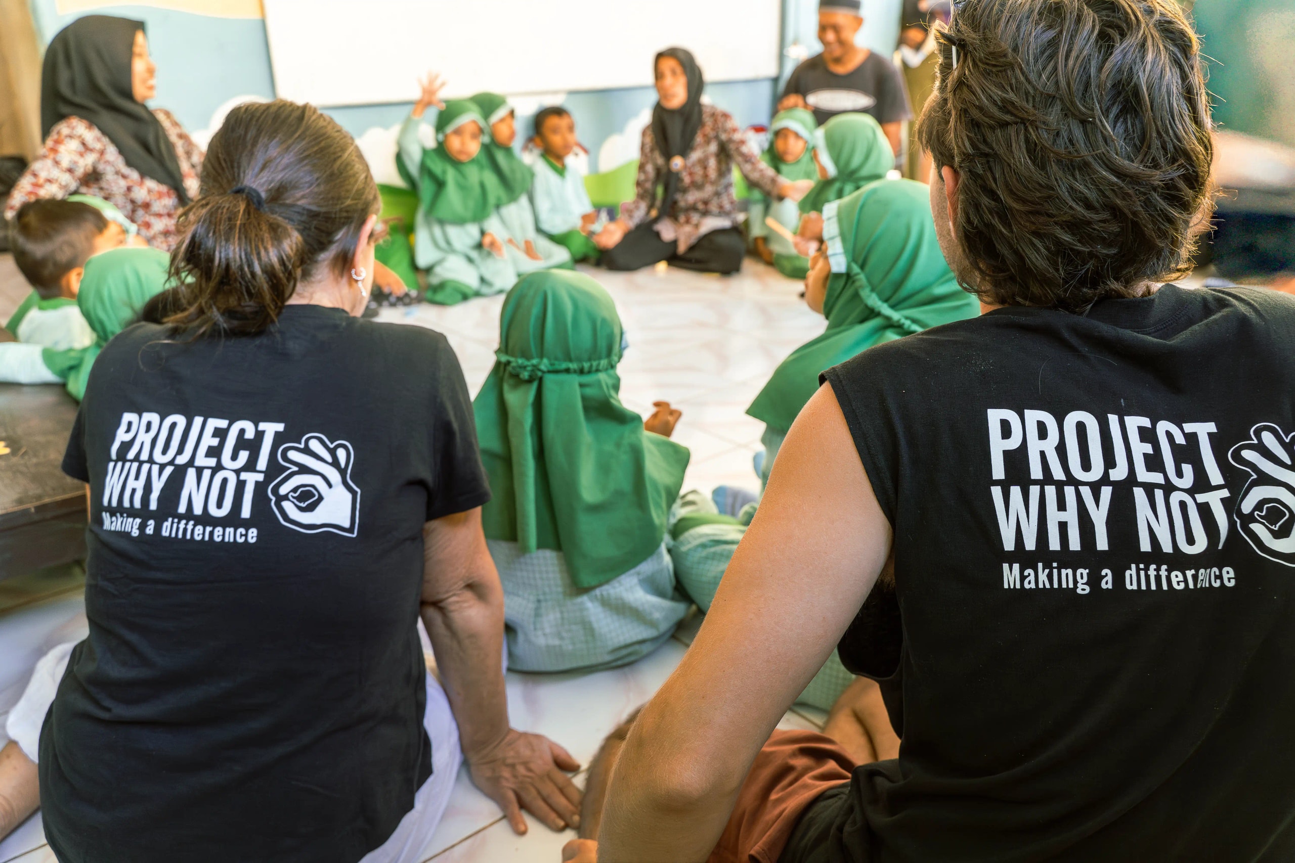 Volunteers wearing "Project Why Not" shirts sitting on the floor with school children and teachers in a classroom setting.