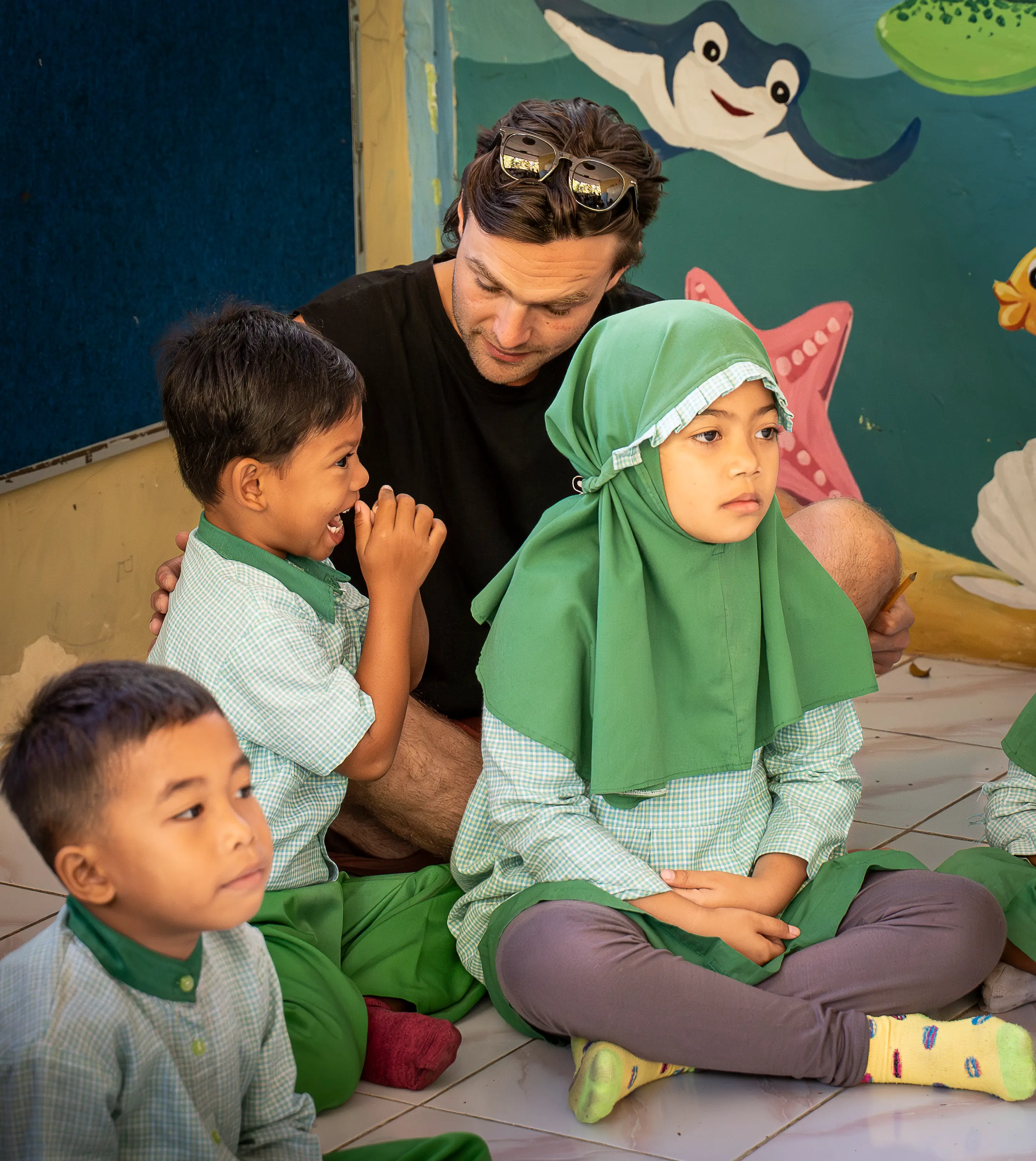 A volunteer sitting on the floor among smiling school children in matching green uniforms inside a classroom.