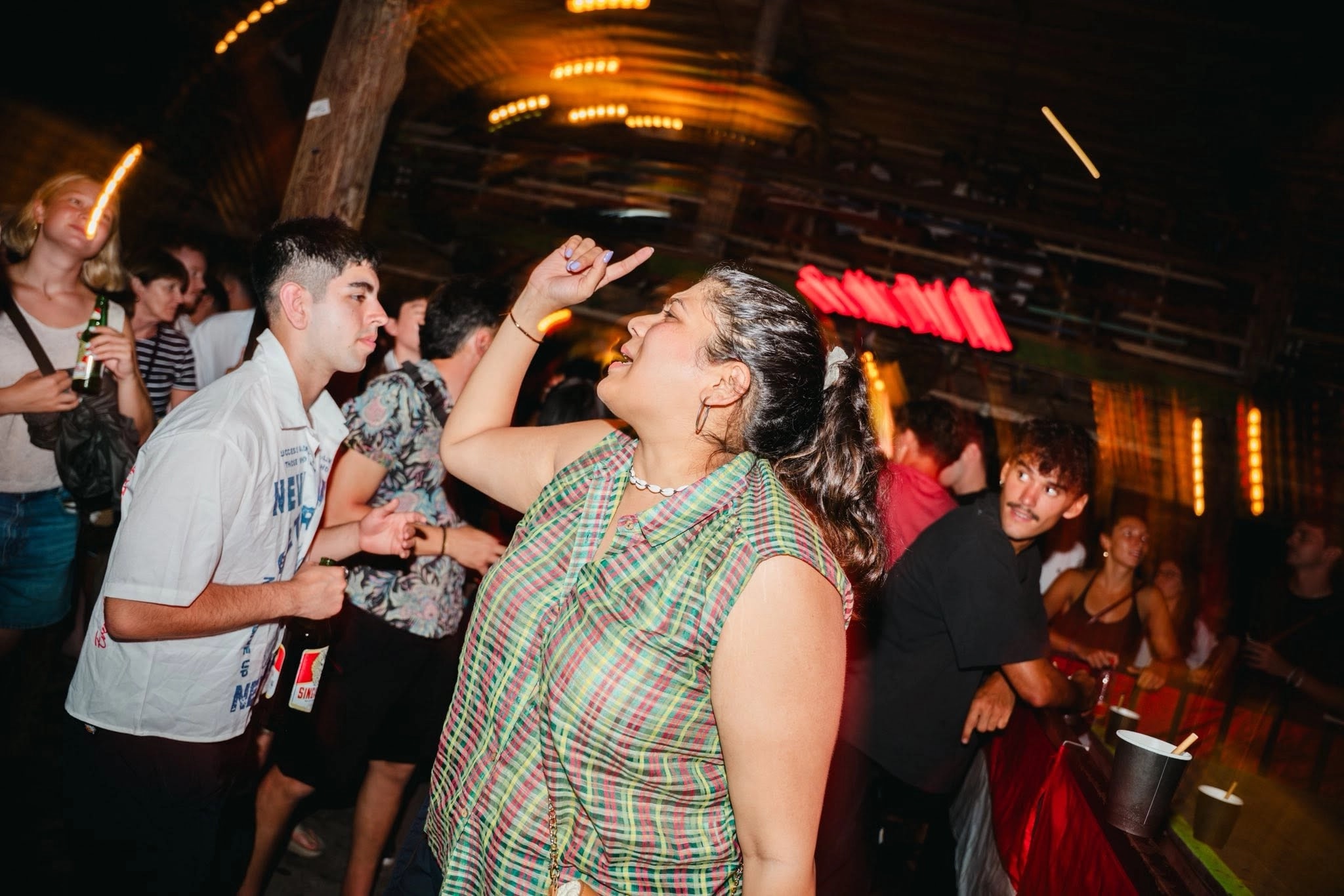 Woman dancing and pointing upward among a lively bar crowd at night.