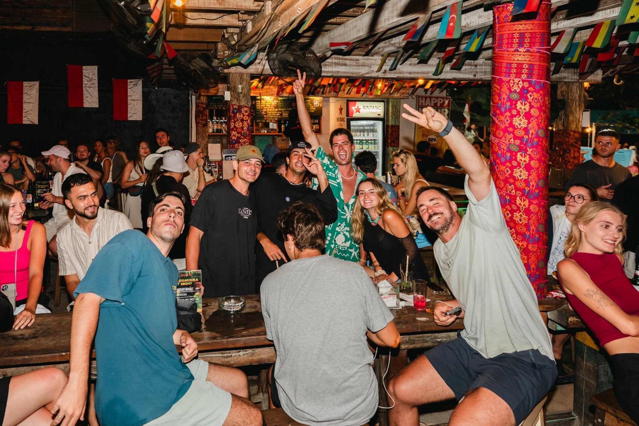 Group of friends sitting around a wooden table flashing peace signs at a beach bar.