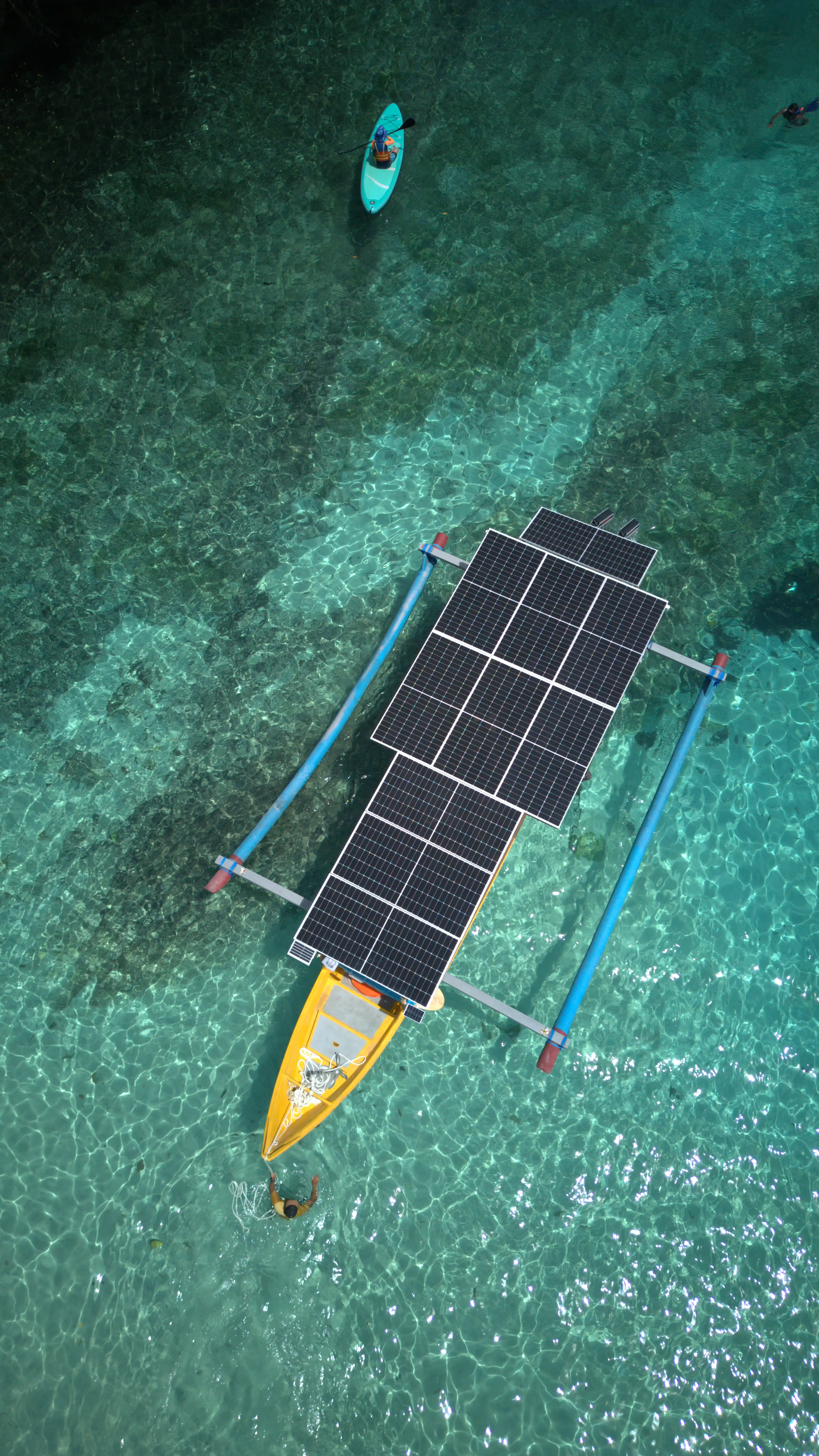 Aerial view of a small yellow outrigger boat fitted with a rooftop solar panel grid in clear tropical water.